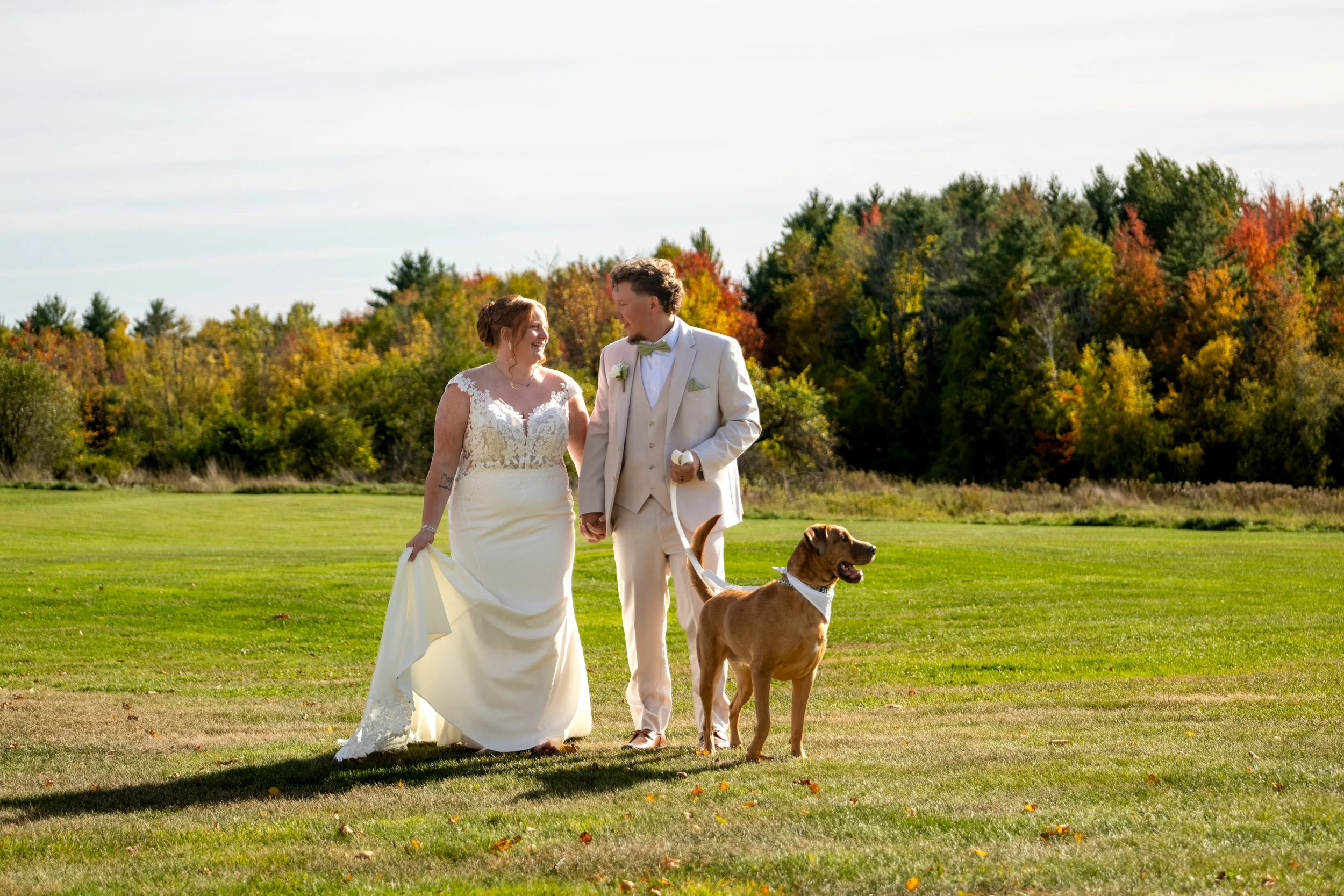 Andrew and Erika walking across the field with Penny beside them and fall trees behind at the Maine Wedding Barn and Event Center in Minot Maine fall wedding photography VMG Productions
