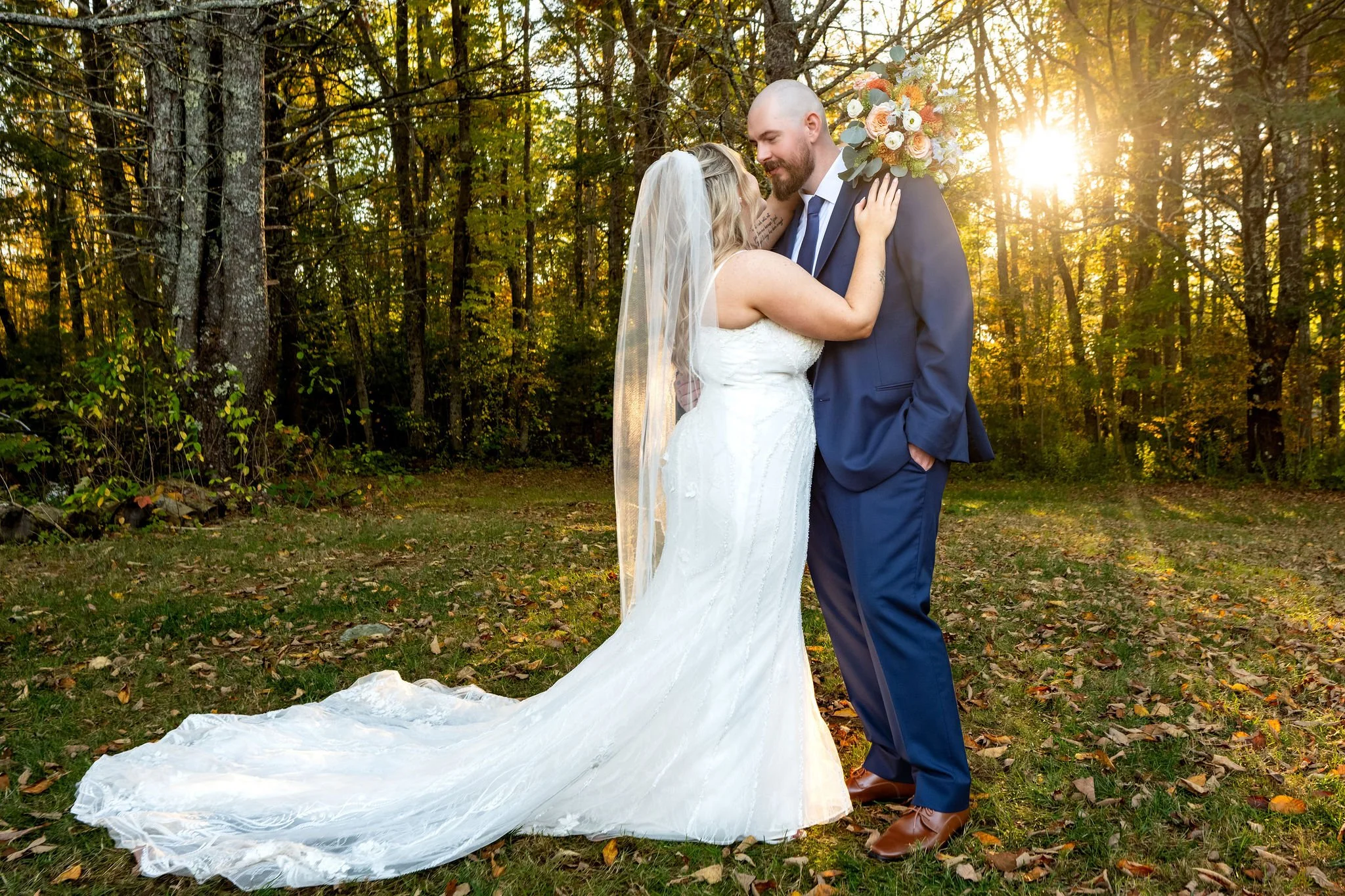 Ellie and Sam embracing in the trees with the low sun flaring through the fall leaves behind them at Boothbay Playhouse in Boothbay Maine fall wedding photography VMG Productions