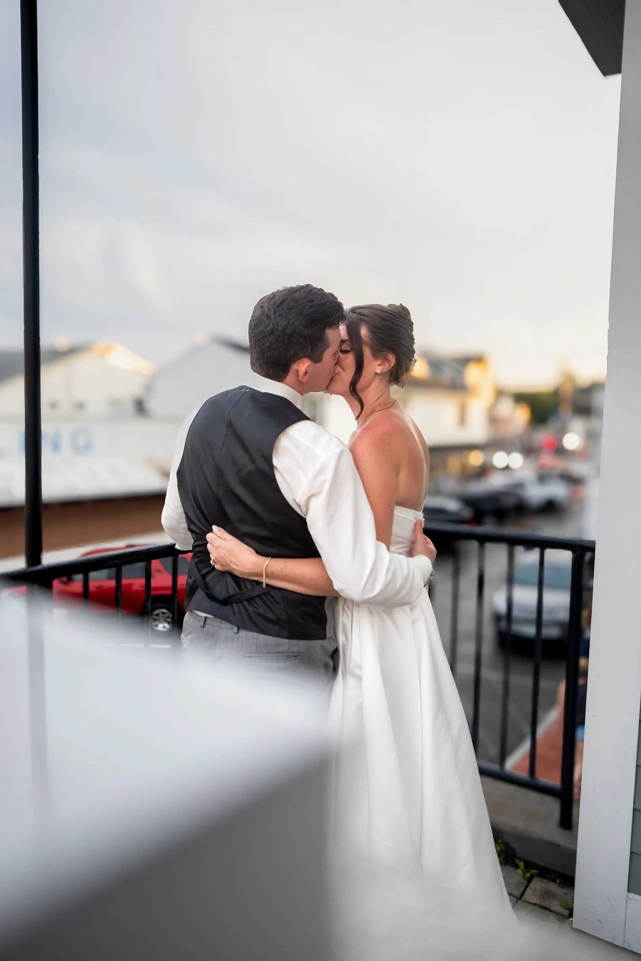 Caroline and Anthony Pirolli kissing on the balcony at dusk with the streets of York Beach below them at Union Bluff Hotel in Maine wedding photography VMG Productions