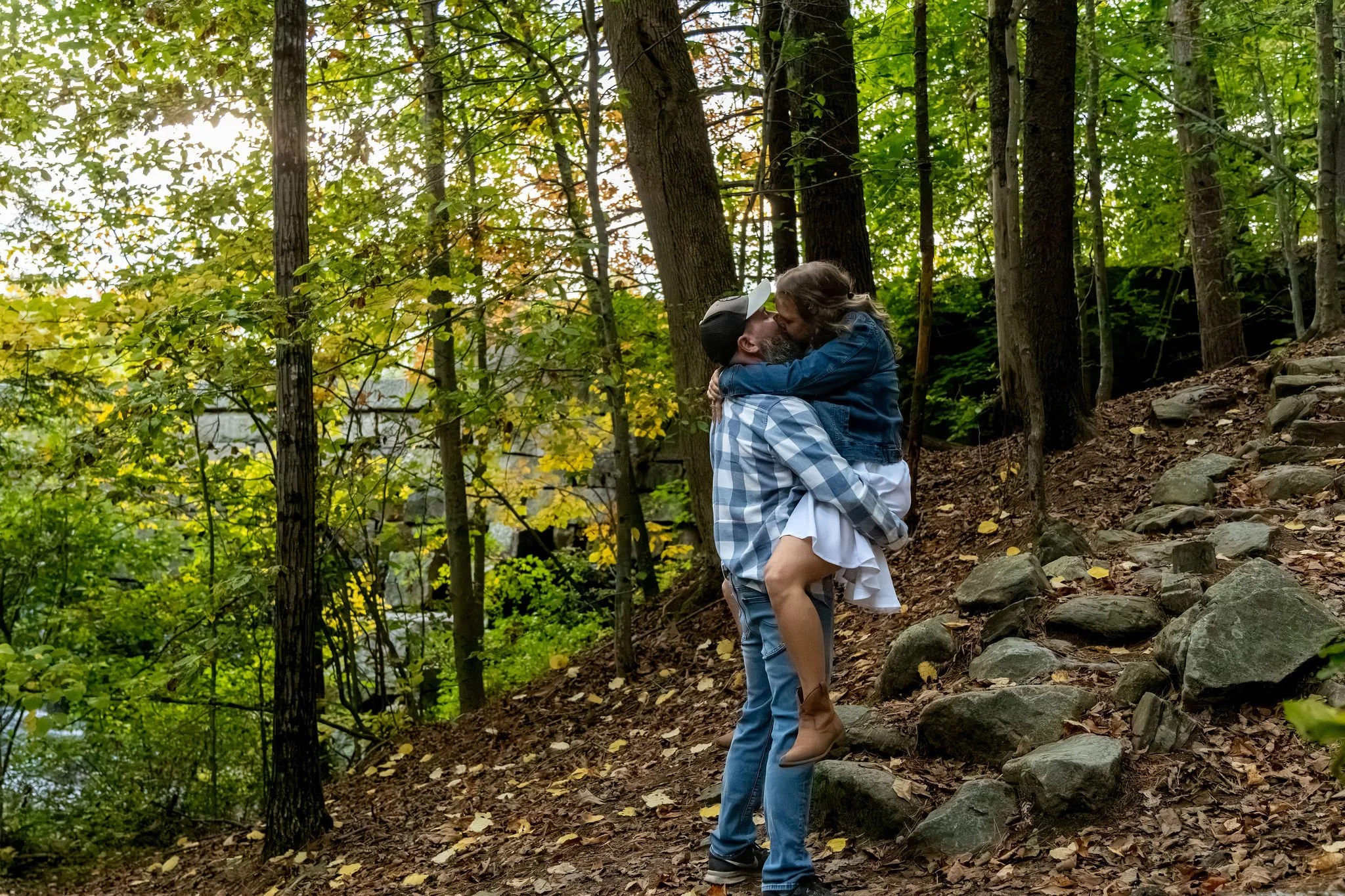 Dave lifting Alison for a kiss on the stone stairs with the low sun coming through the trees at Vaughan Woods in Hallowell Maine fall engagement photography VMG Productions