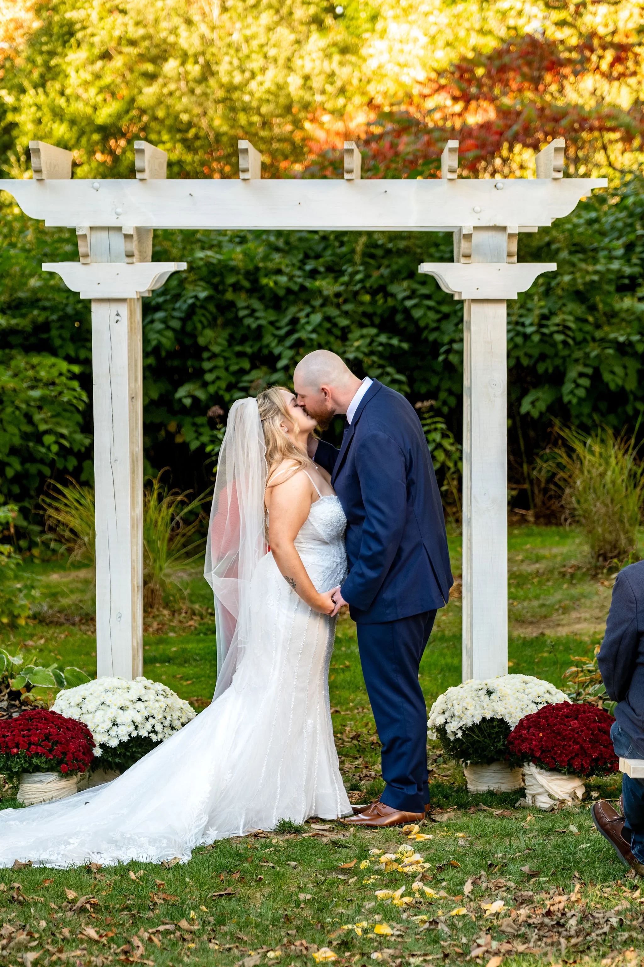 Ellie and Sam sharing their first kiss as a married couple under the white pergola with mums at their feet and fall foliage behind at Boothbay Playhouse in Boothbay Maine fall wedding photography VMG Productions