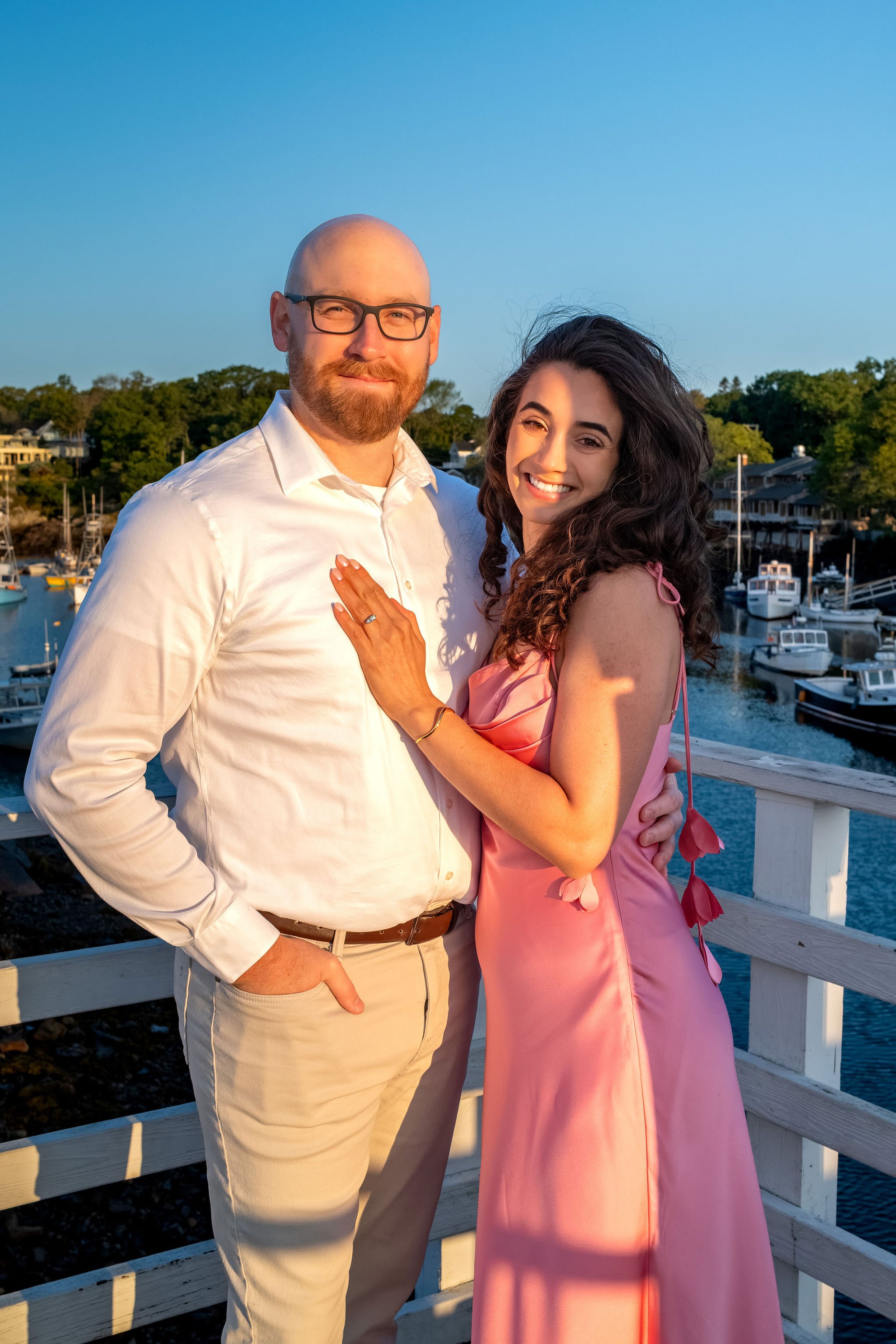 Zack and Helena smiling at the camera on the footbridge with the harbor and boats behind them on Marginal Way in Ogunquit Maine sunrise proposal and engagement photography VMG Productions