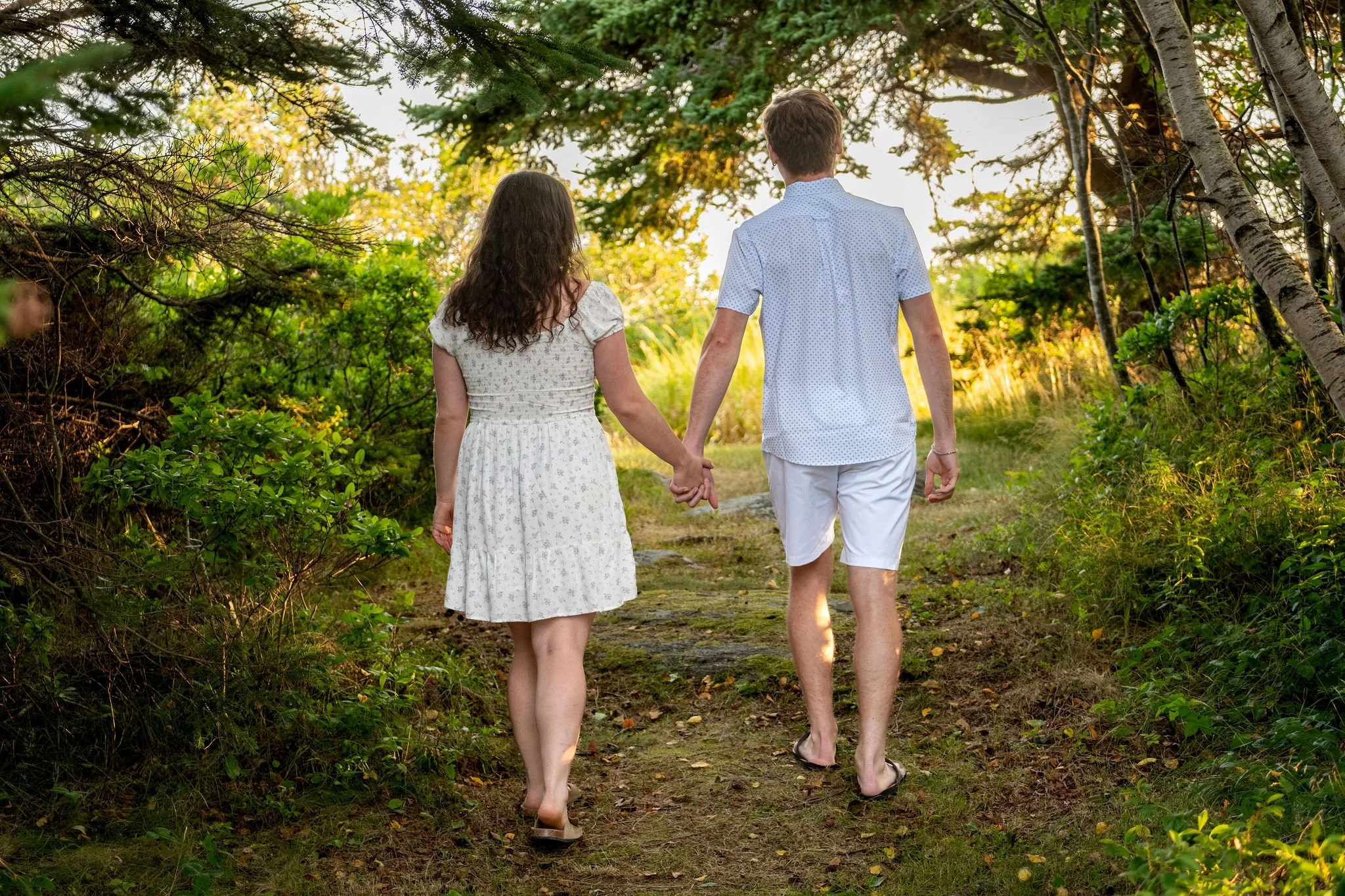 Kirstie and Lucas walking hand in hand away from the camera down a wooded path at Reid State Park in Georgetown Maine engagement photography VMG Productions