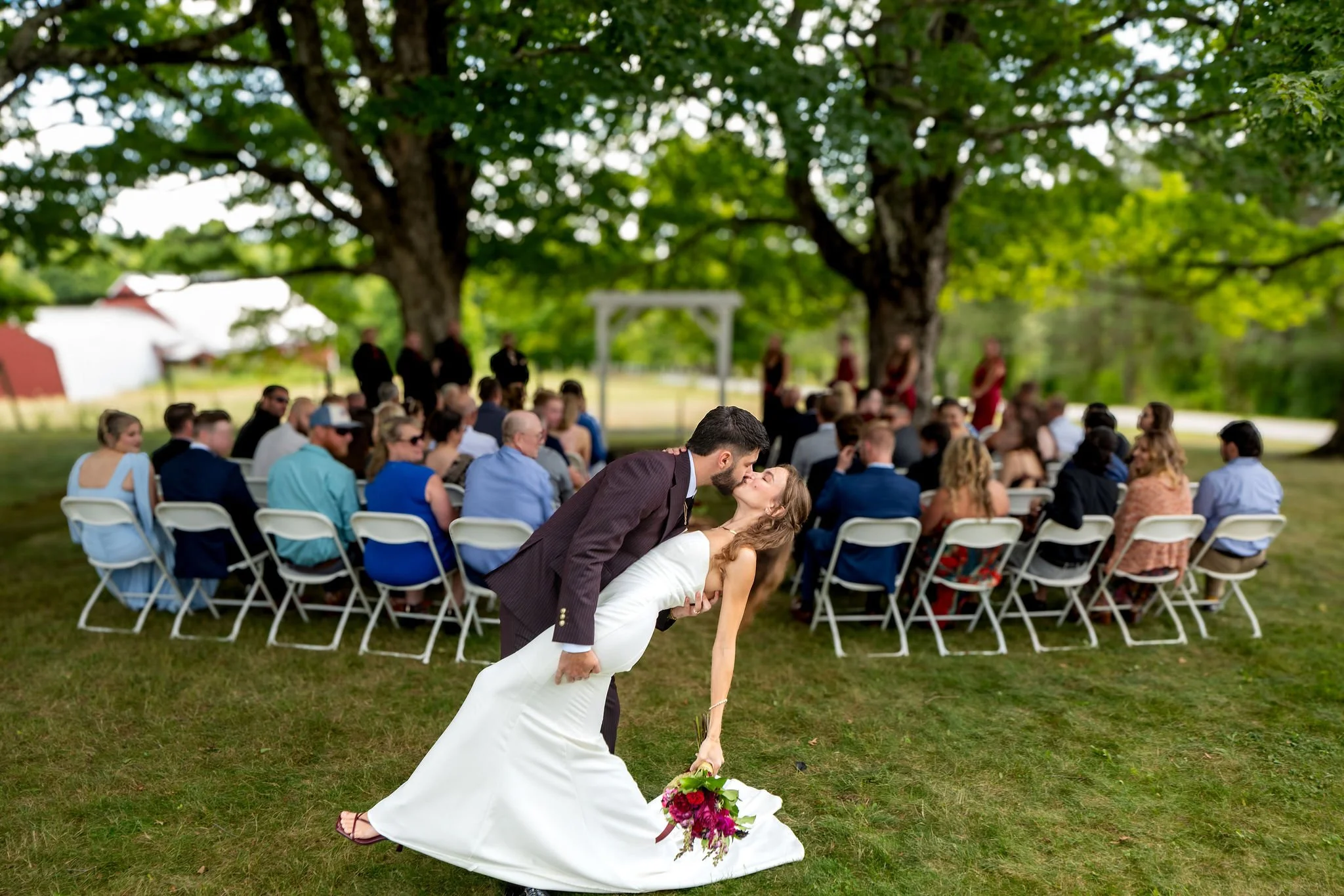 Cassidy and Chase Begin dip kiss in front of their seated guests under the maples at Maple Hill Farm Inn and Conference Center in Hallowell Maine summer wedding photography VMG Productions