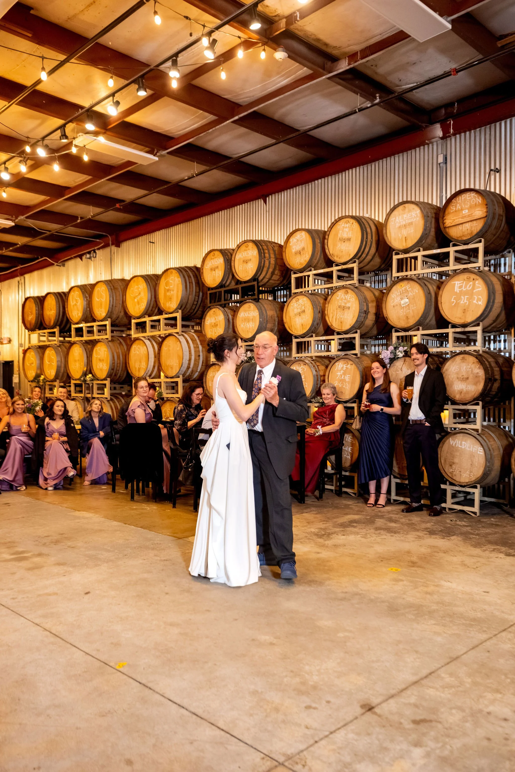 Victoria dancing with her dad in front of the stacked barrels as guests watch at The Cellars at Allagash Brewing Company in Portland Maine fall wedding photography VMG Productions