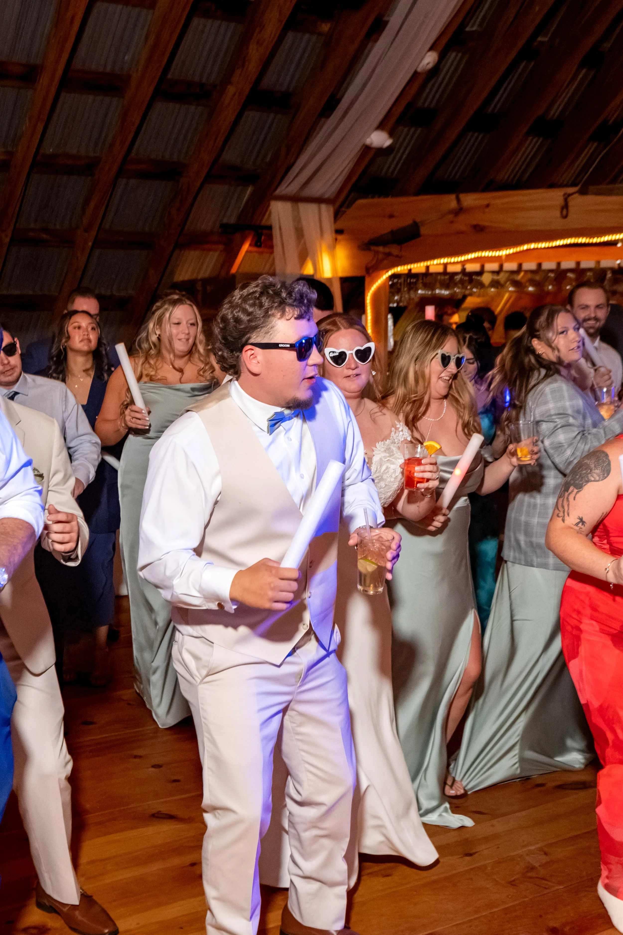 A groomsman in a vest and sunglasses dancing with a light-up wand in the middle of the crowd at the Maine Wedding Barn and Event Center in Minot Maine fall wedding photography VMG Productions