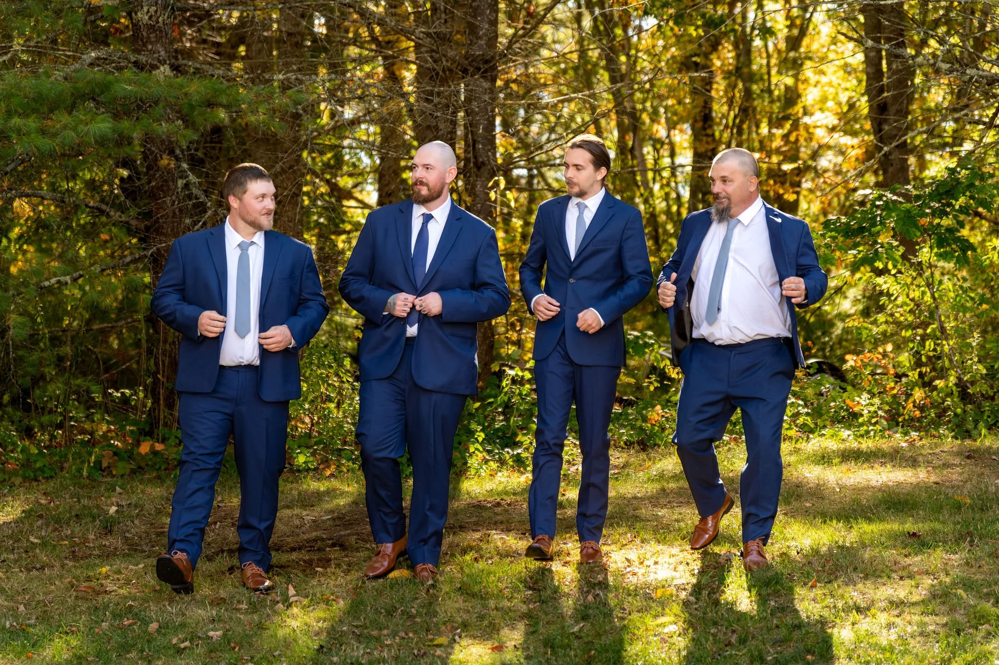 Sam and his groomsmen in navy suits walking together across the grass with the fall woods behind them at Boothbay Playhouse in Boothbay Maine fall wedding photography VMG Productions