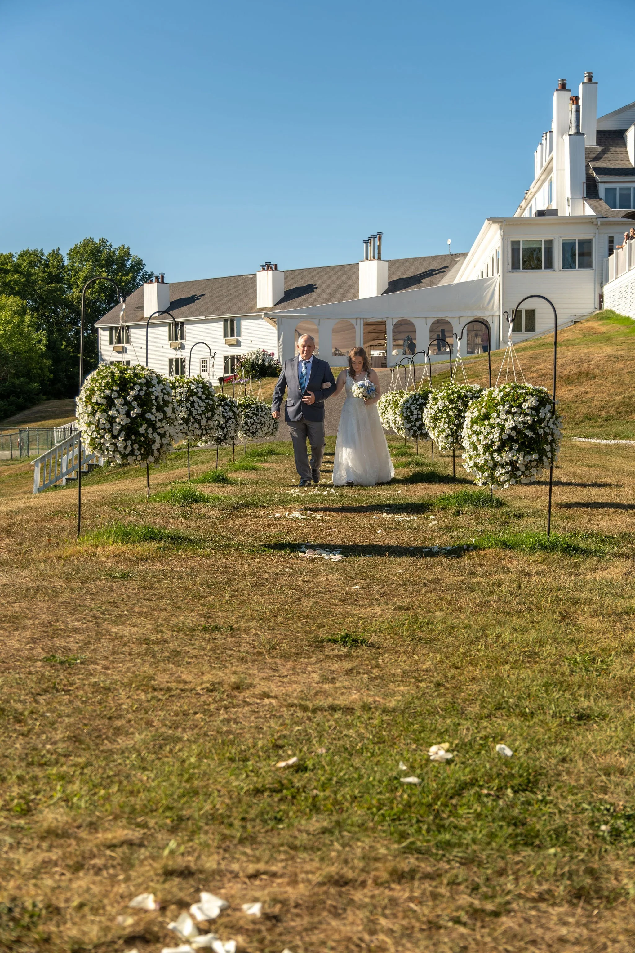 Hollie walking down the hillside aisle on her dad's arm with the white inn on the ridge behind them at The Lucerne Inn in Dedham Maine summer wedding photography VMG Productions