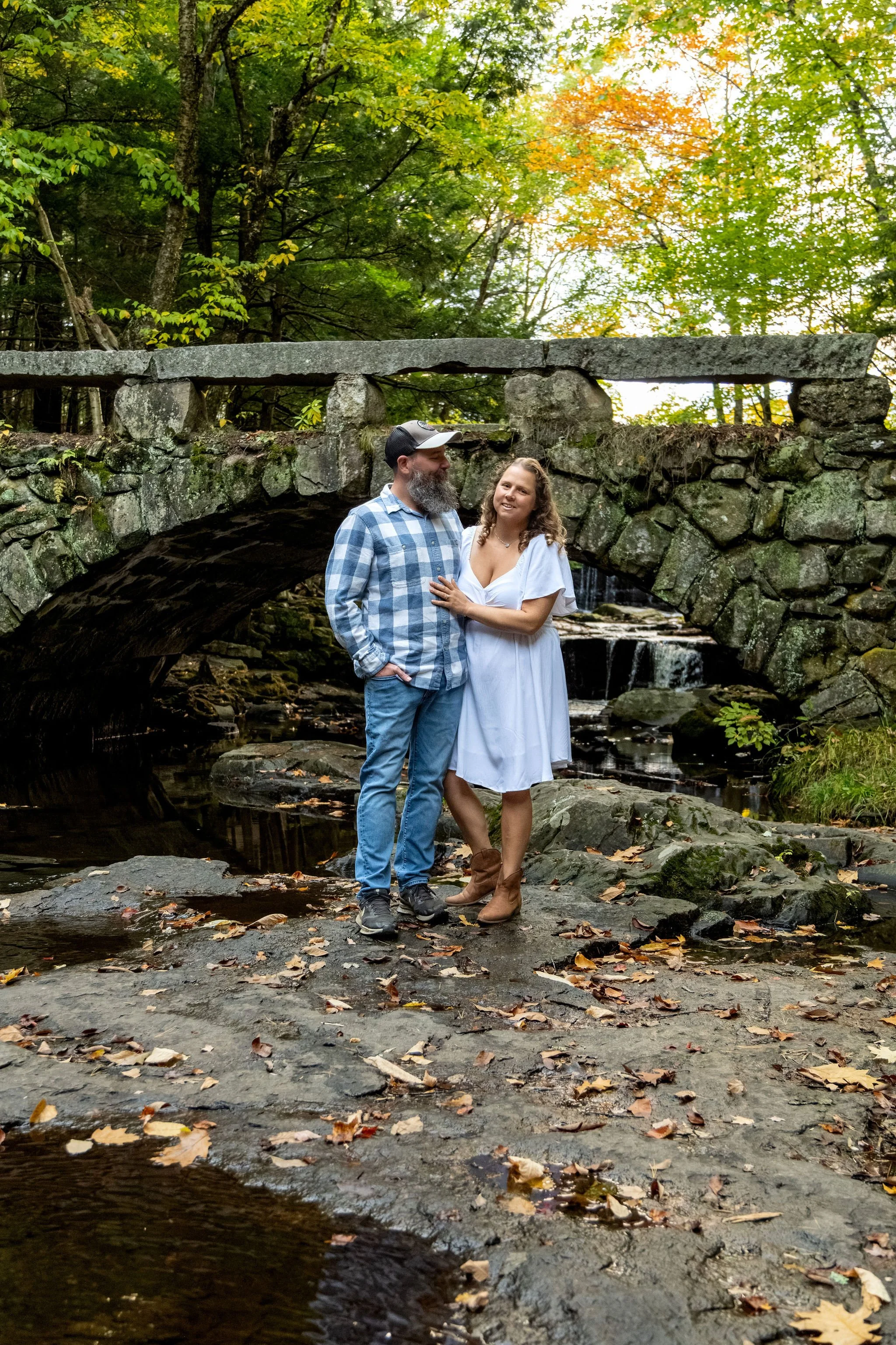 Alison and Dave standing below the arch bridge with fallen leaves scattered across the wet ledge and orange maples overhead at Vaughan Woods in Hallowell Maine fall engagement photography VMG Productions
