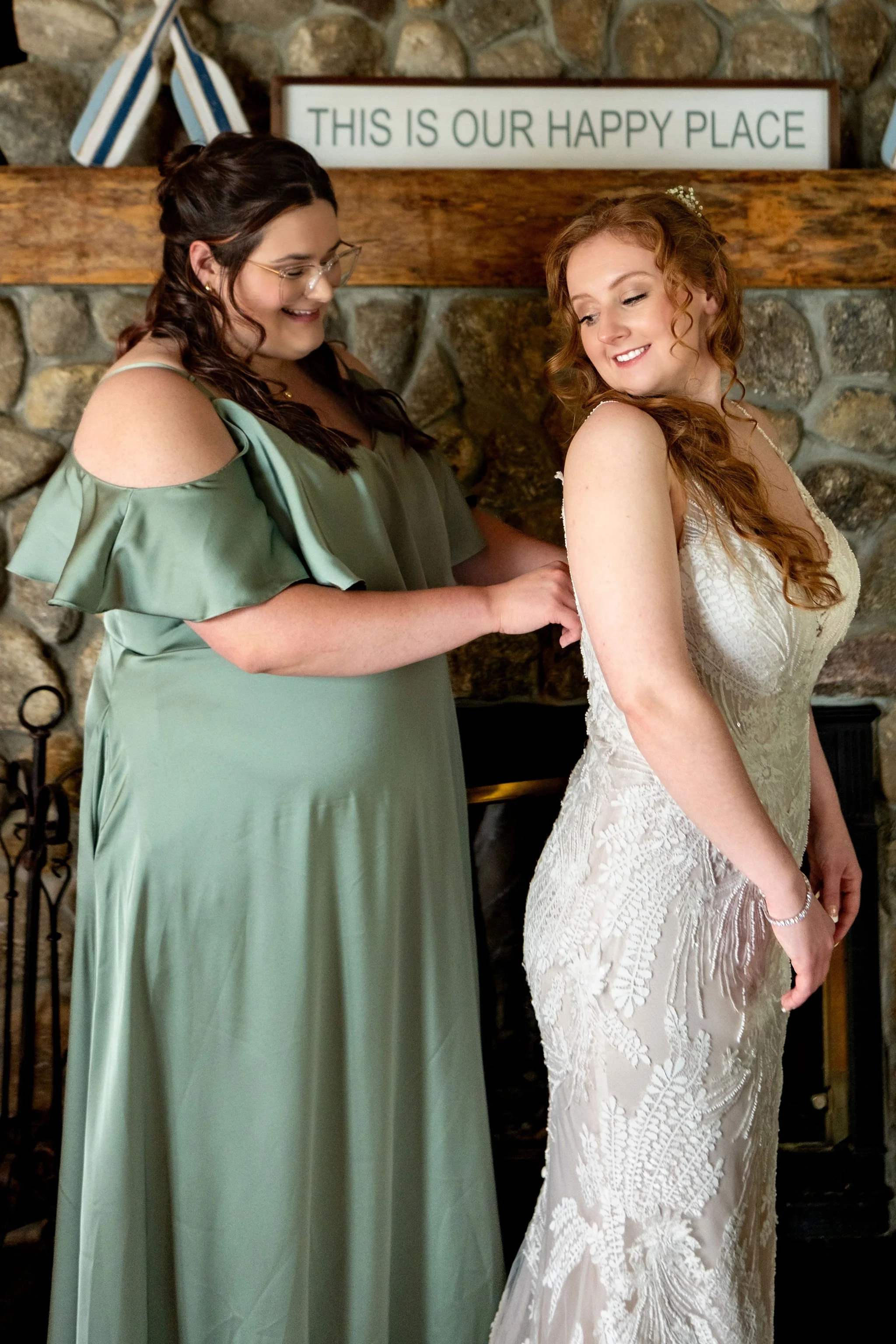 Kayla DeAngelis having her gown fastened by her bridesmaid in front of the stone fireplace at Lake Winnisquam Belmont New Hampshire summer wedding photography VMG Productions