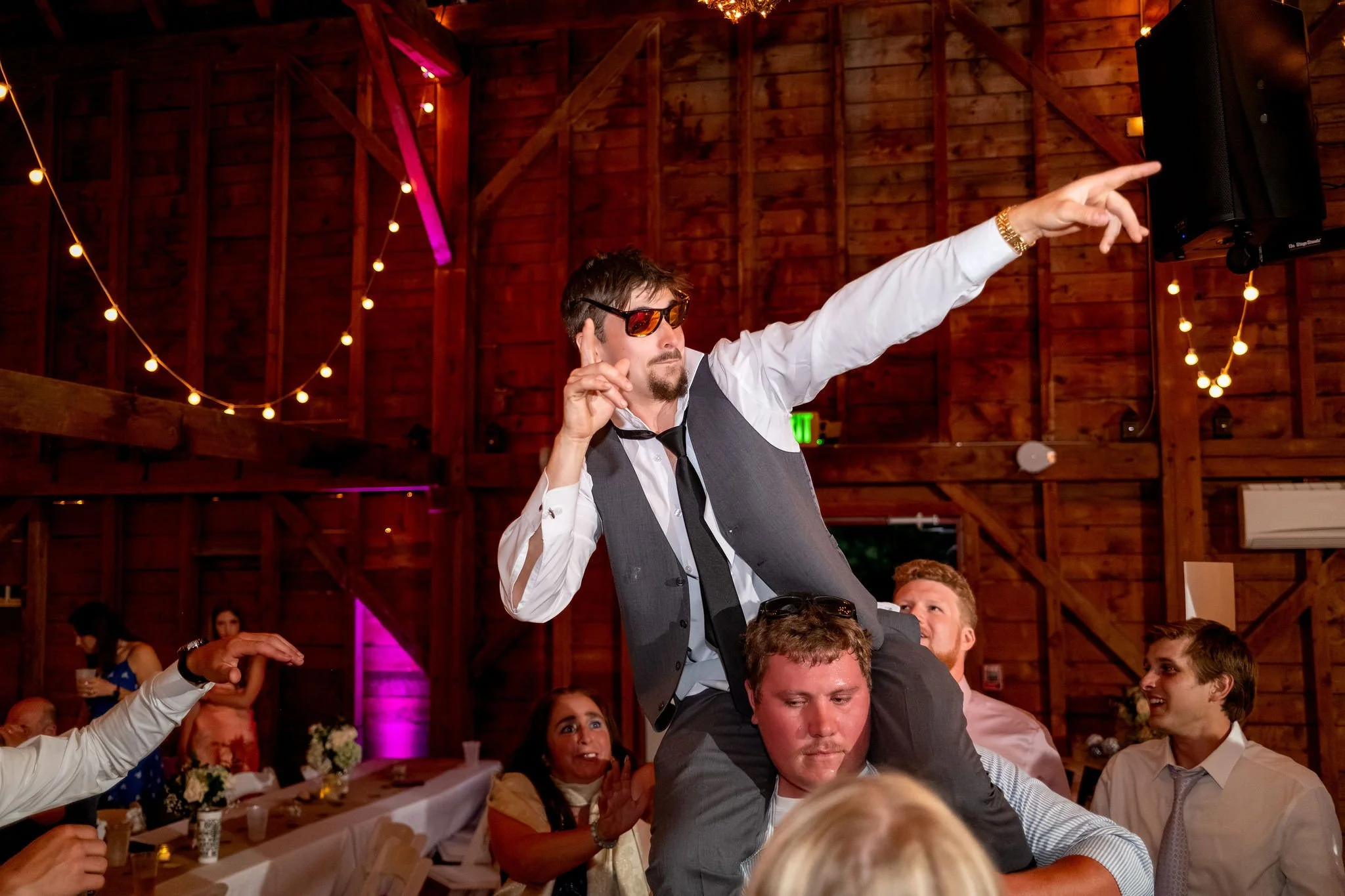 Alyssa and Colby a guest riding on shoulders above the dance floor pointing across the barn at the 1888 Barn Bethel Maine summer wedding photography VMG Productions