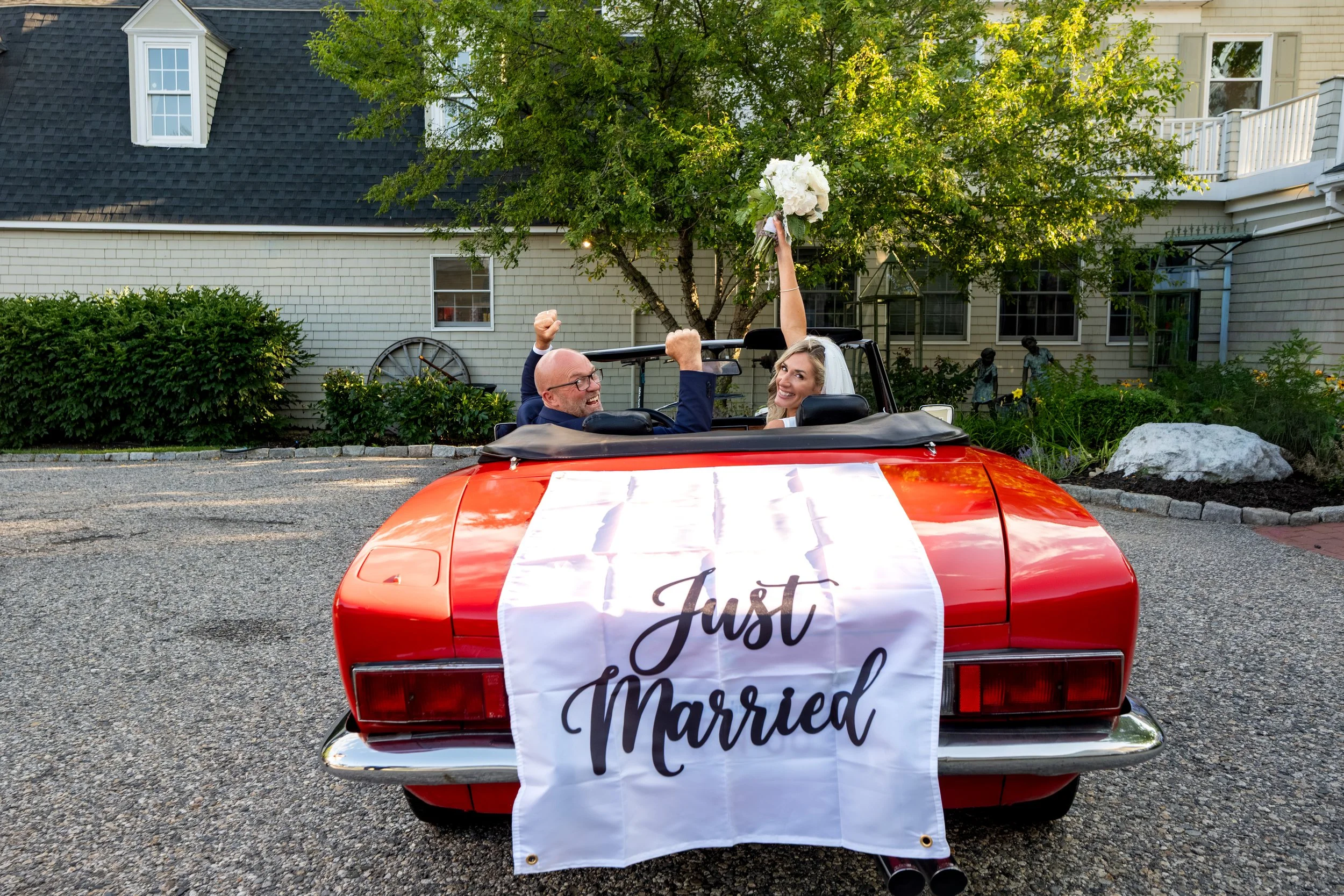Ashley and Marc Draper celebrating with fists and bouquet raised behind the Just Married sign on the vintage red Fiat at The Breakwater Inn in Kennebunkport Maine summer wedding photography VMG Productions