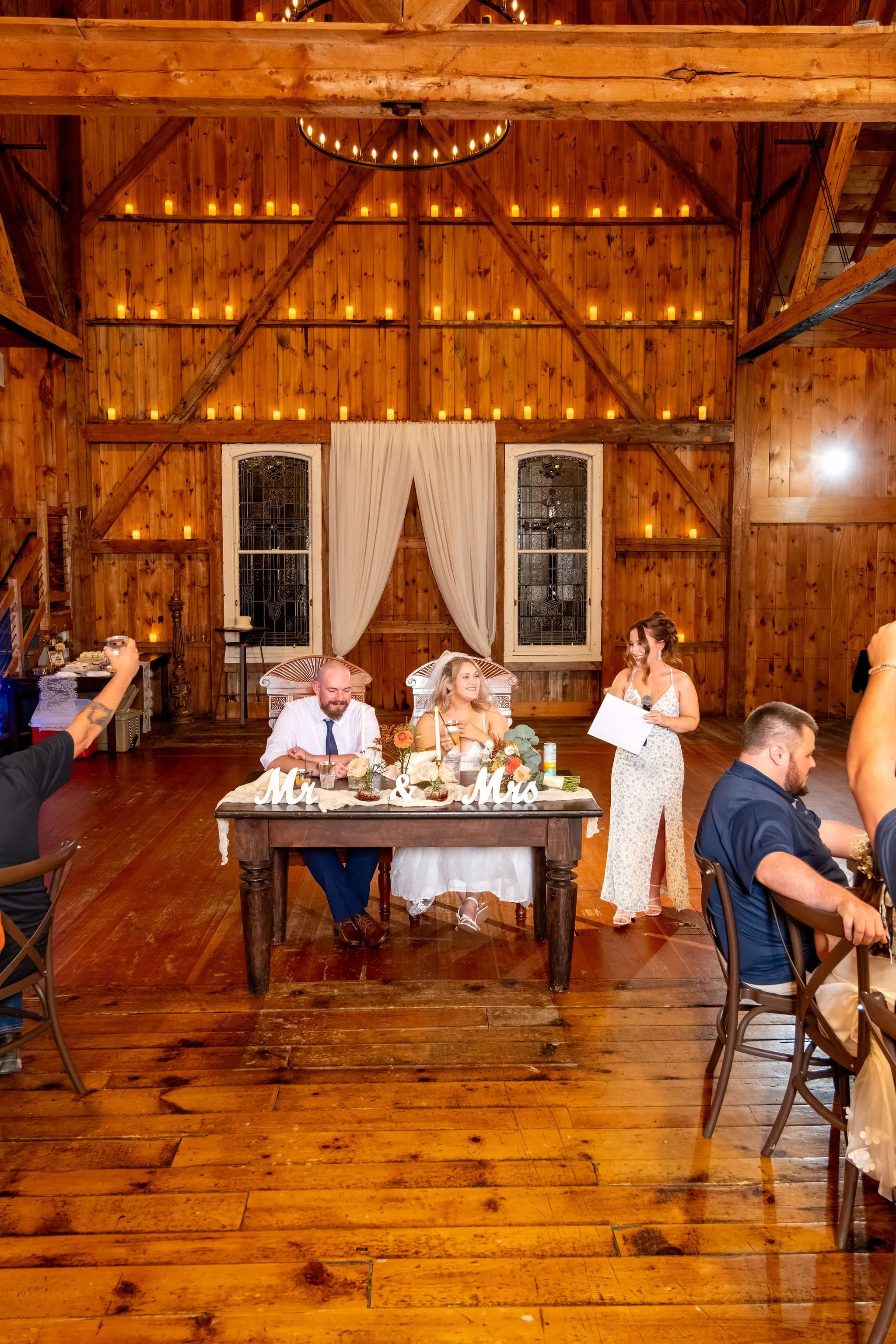 Wide view of the barn as the maid of honor gives her toast with Ellie and Sam listening at the sweetheart table at Boothbay Playhouse in Boothbay Maine fall wedding photography VMG Productions