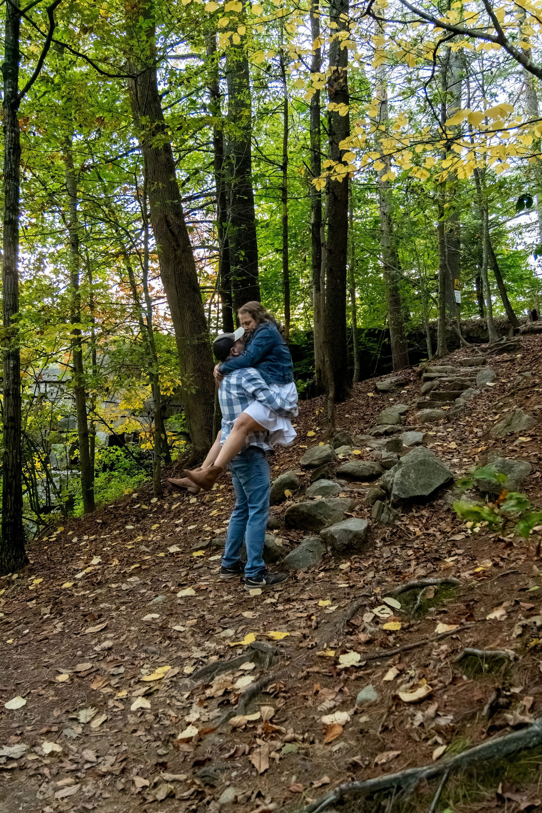 Dave lifting Alison off the stone steps with the yellow woods around them at Vaughan Woods in Hallowell Maine fall engagement photography VMG Productions