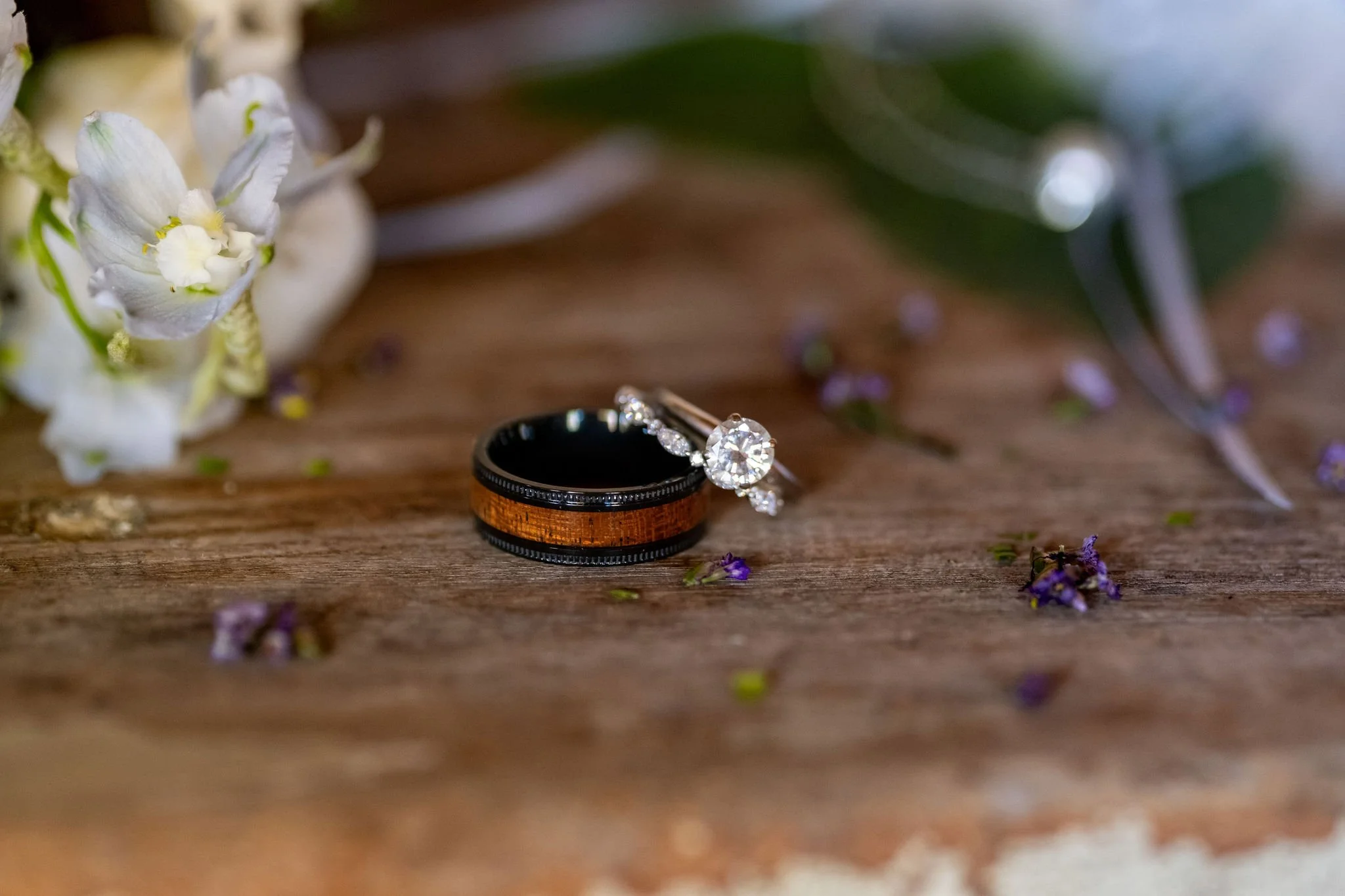 Wedding rings resting on weathered wood among white flowers and tiny purple petals at Boothbay Playhouse in Boothbay Maine fall wedding photography VMG Productions