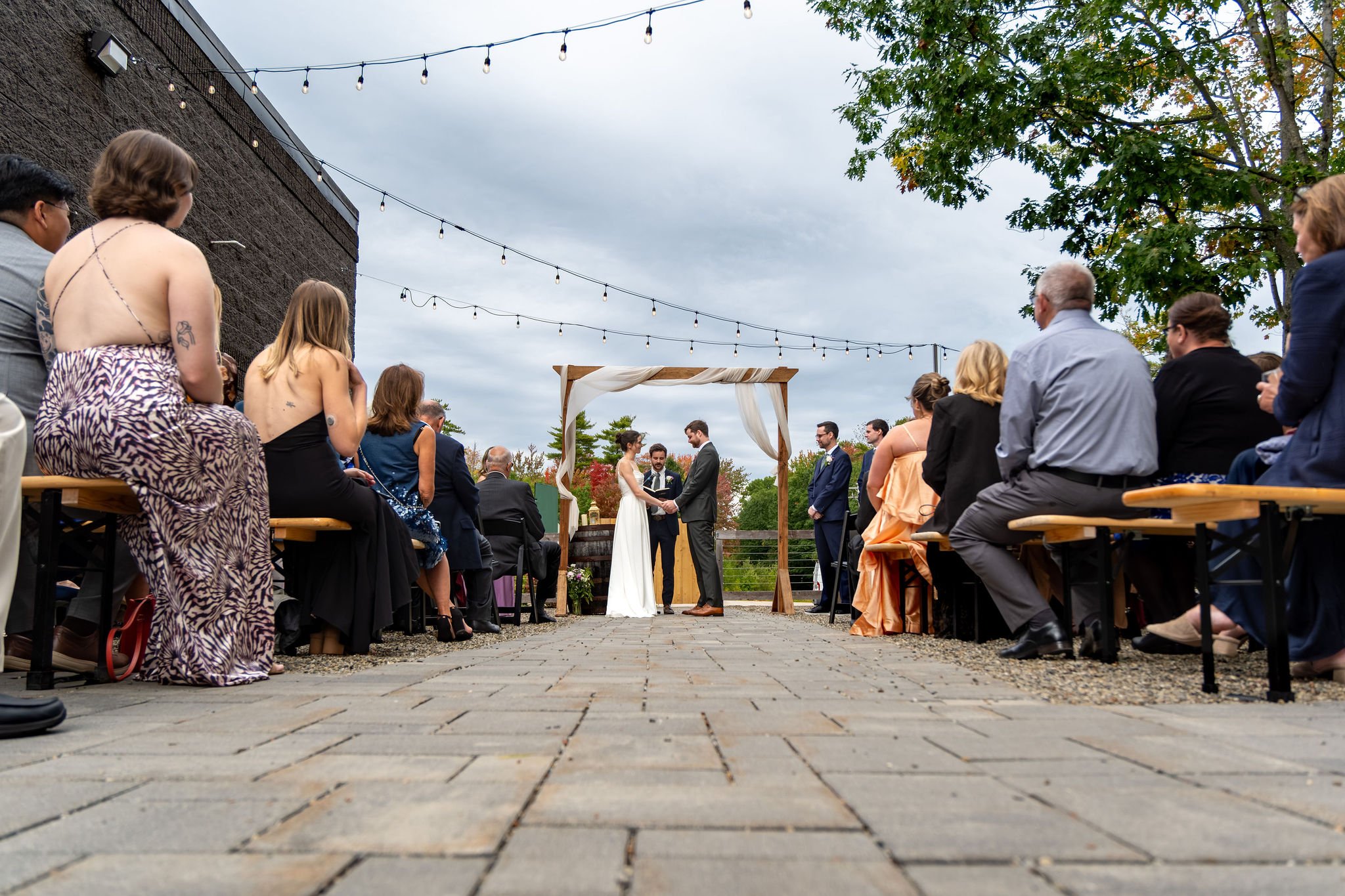 Wide view of the courtyard ceremony from the back of the aisle with string lights and the wooden arbor at The Cellars at Allagash Brewing Company in Portland Maine fall wedding photography VMG Productions