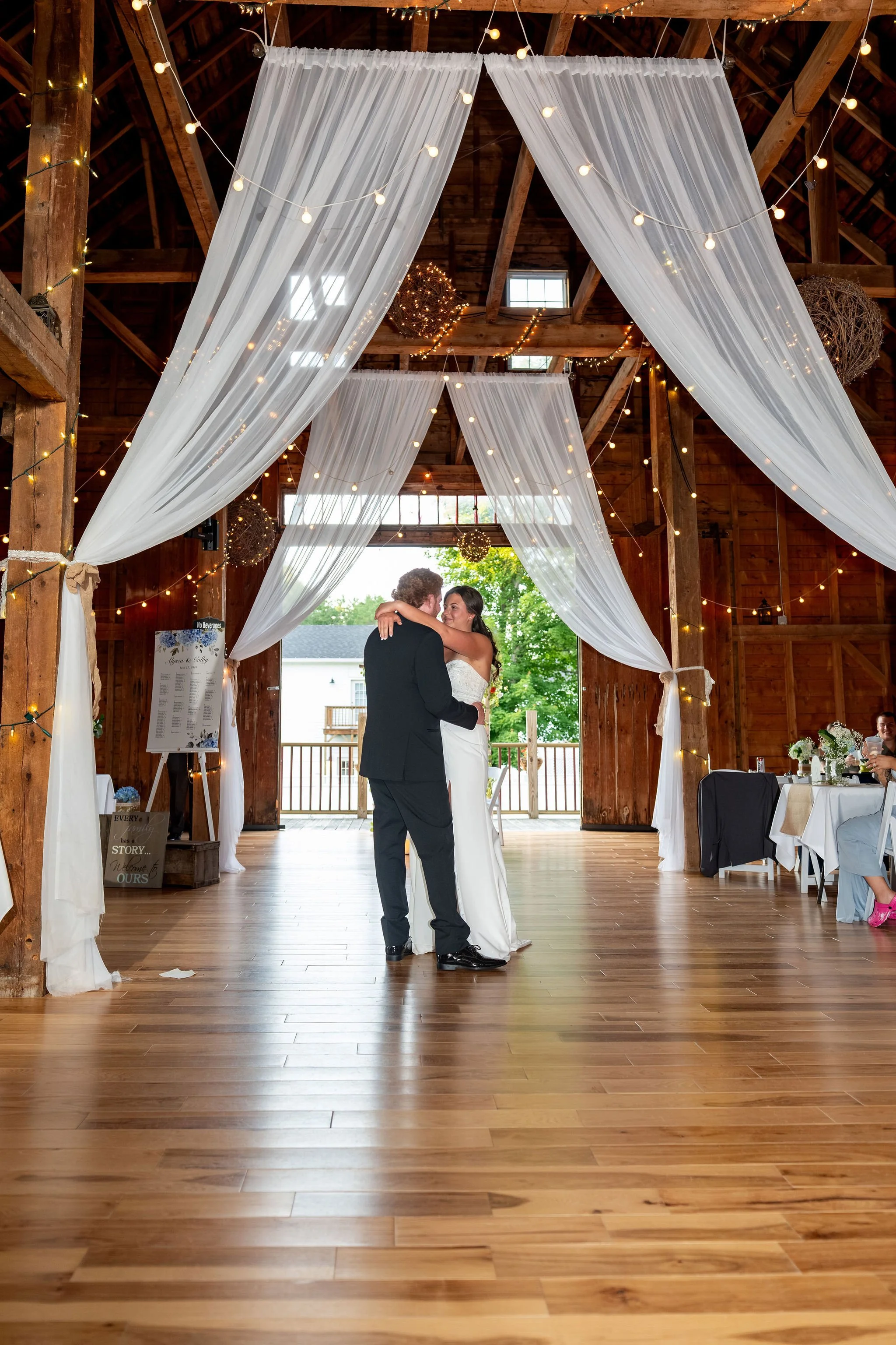 Alyssa and Colby sharing their first dance beneath white draping and string lights at the 1888 Barn Bethel Maine summer wedding photography VMG Productions