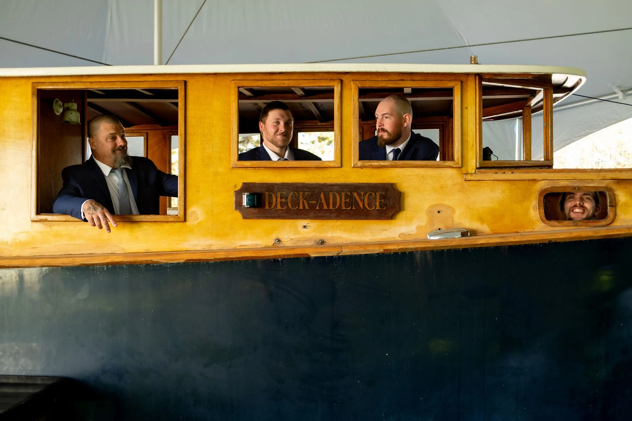 Three groomsmen leaning out the windows of a yellow wooden boat with one poking his head through a porthole at Boothbay Playhouse in Boothbay Maine fall wedding photography VMG Productions