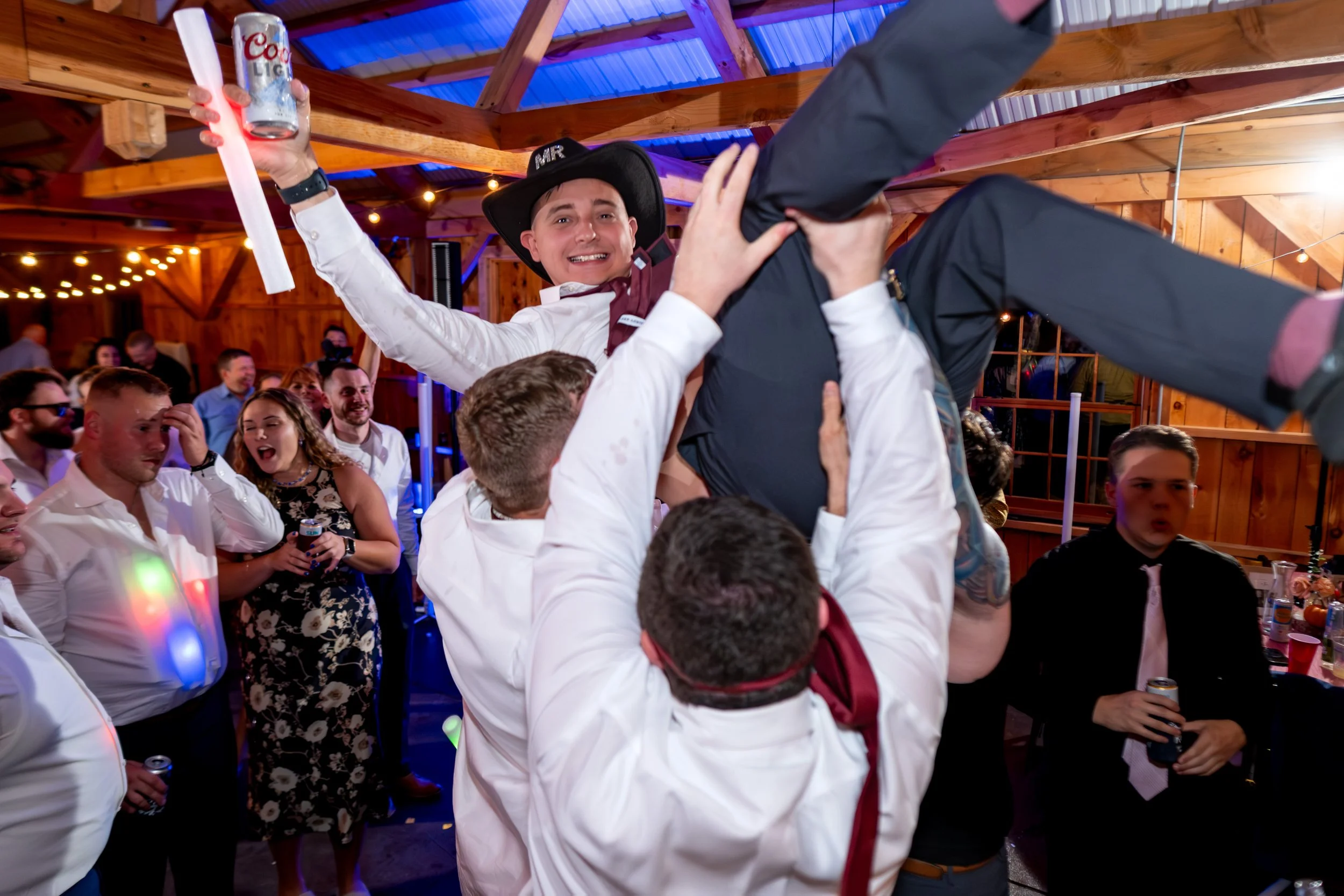 Groom wearing MR cowboy hat being lifted by groomsmen on full dance floor at Sites Farm Athens Maine rustic fall barn wedding reception 2025 with guests cheering and celebrating with colorful LED glow sticks under blue barn rafters