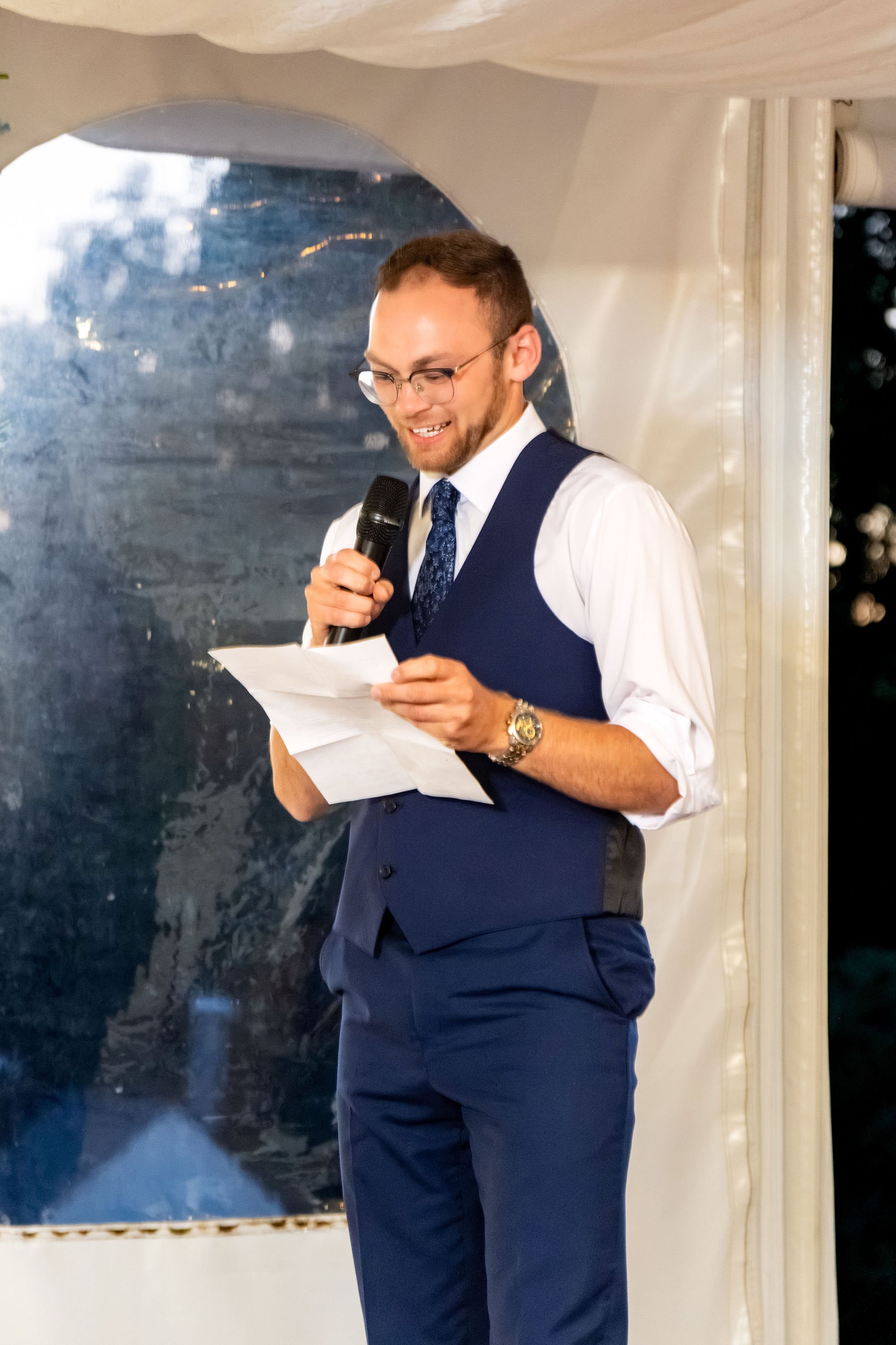 The best man reading his speech from a folded page under the tent at The Lucerne Inn in Dedham Maine summer wedding photography VMG Productions