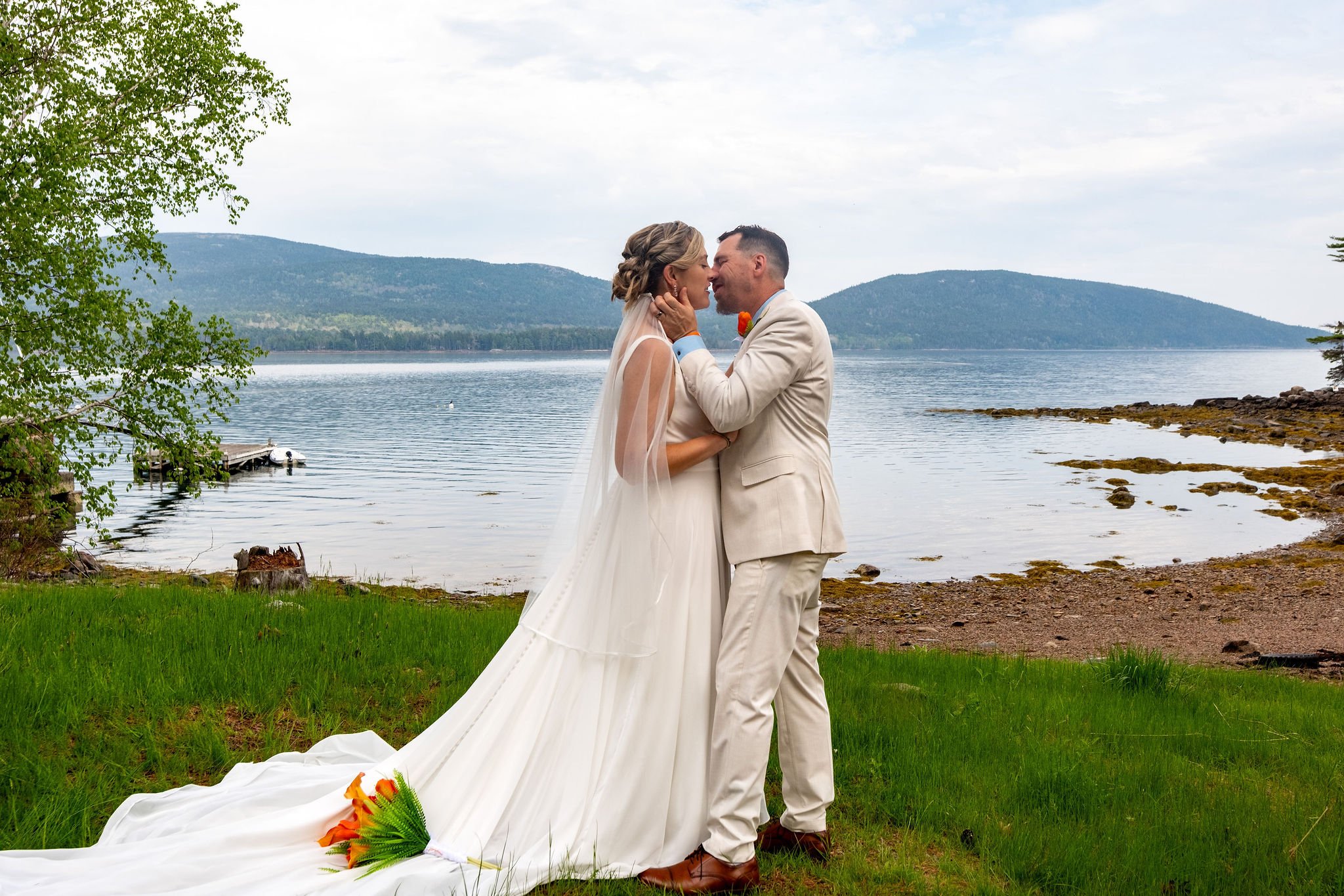 Alli and Adam Baudoin kissing on the dock with the full sweep of Somes Sound and the Acadia hills behind them in Mount Desert Maine spring wedding photography VMG Productions