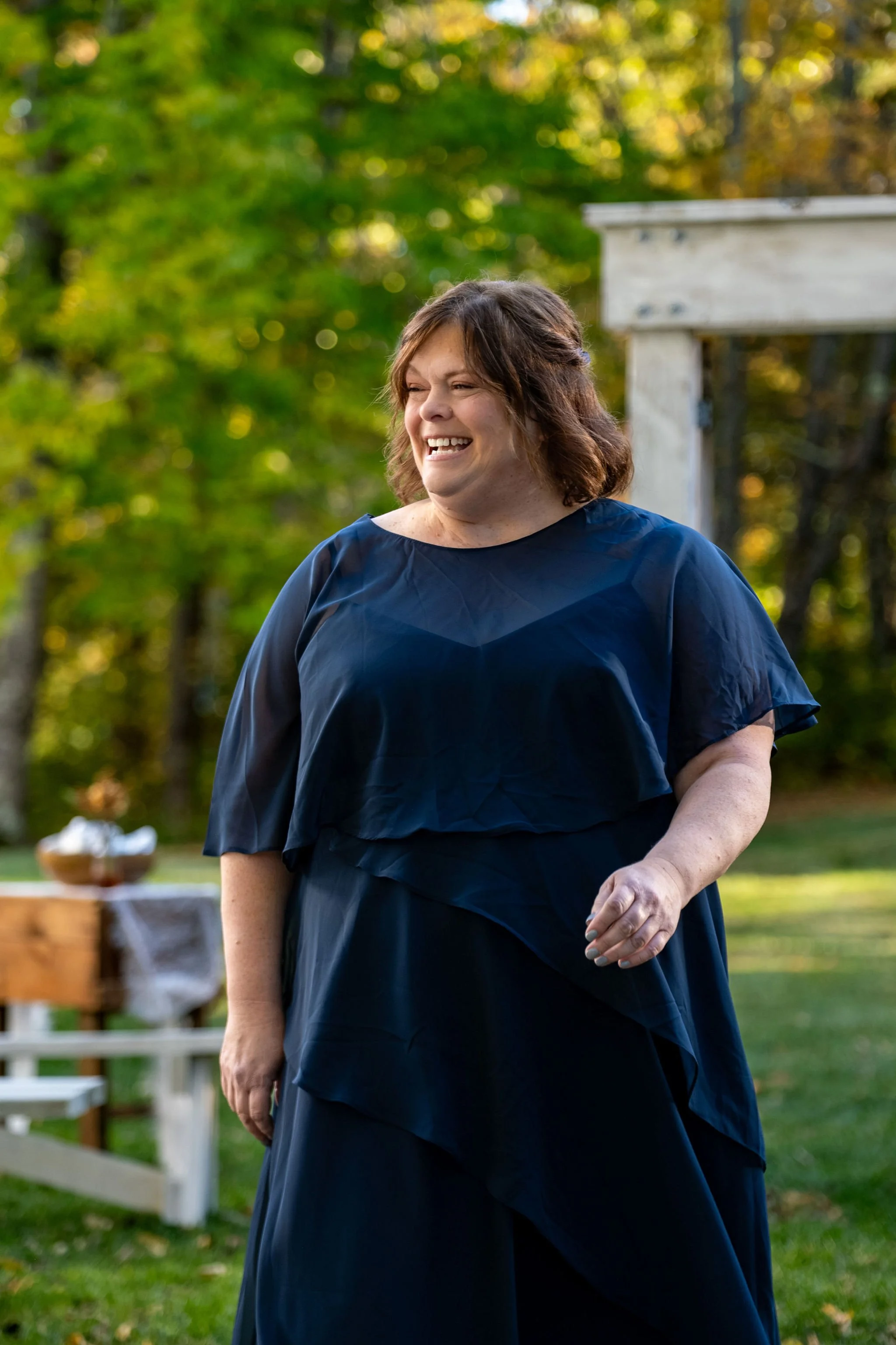 Ellie's mom in a navy dress smiling as she waits on the lawn before the ceremony at Boothbay Playhouse in Boothbay Maine fall wedding photography VMG Productions