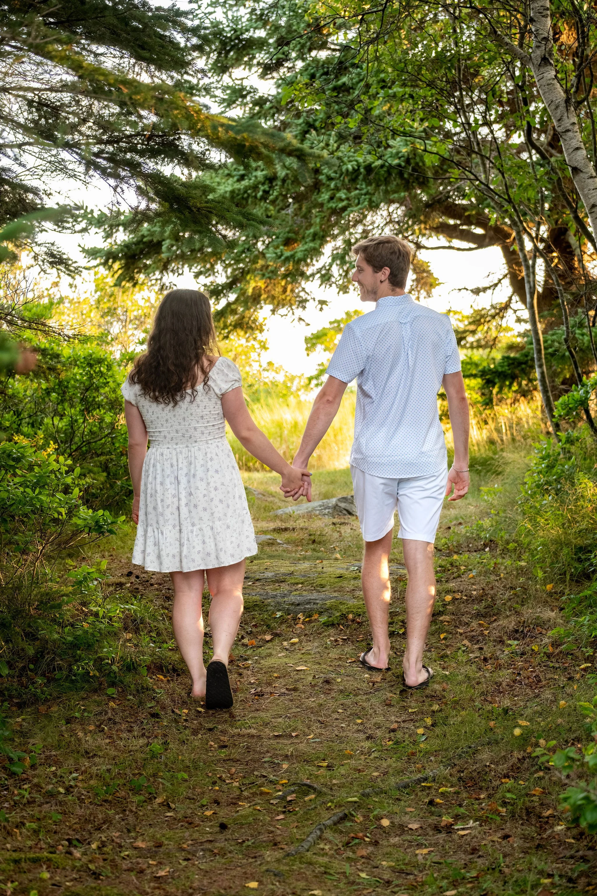 Kirstie and Lucas holding hands on a sunlit path through the birches and spruce at Reid State Park in Georgetown Maine engagement photography VMG Productions