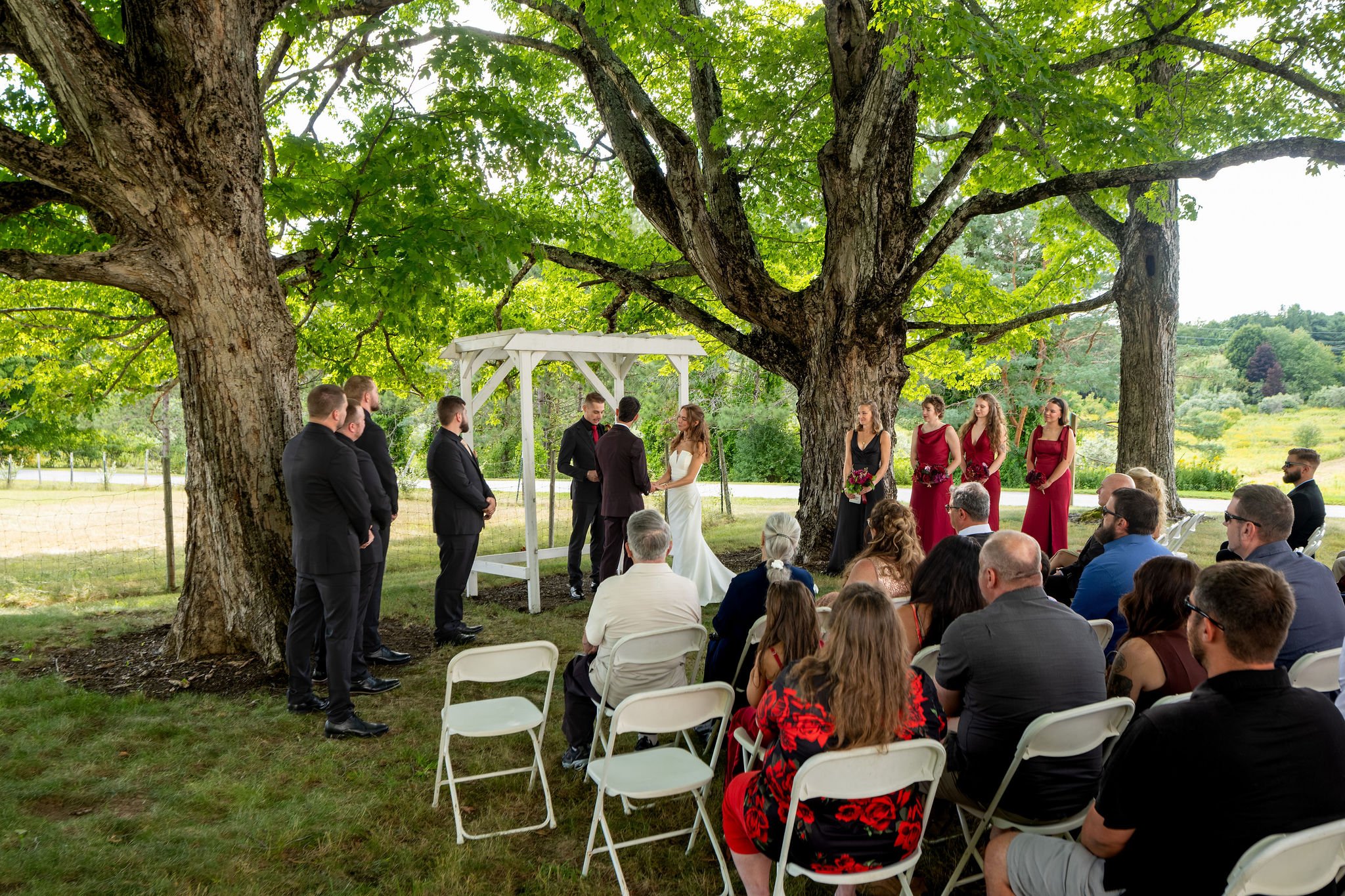 Wide view of Cassidy and Chase Begin's ceremony under the maple trees with guests seated on the lawn at Maple Hill Farm Inn and Conference Center in Hallowell Maine summer wedding photography VMG Productions
