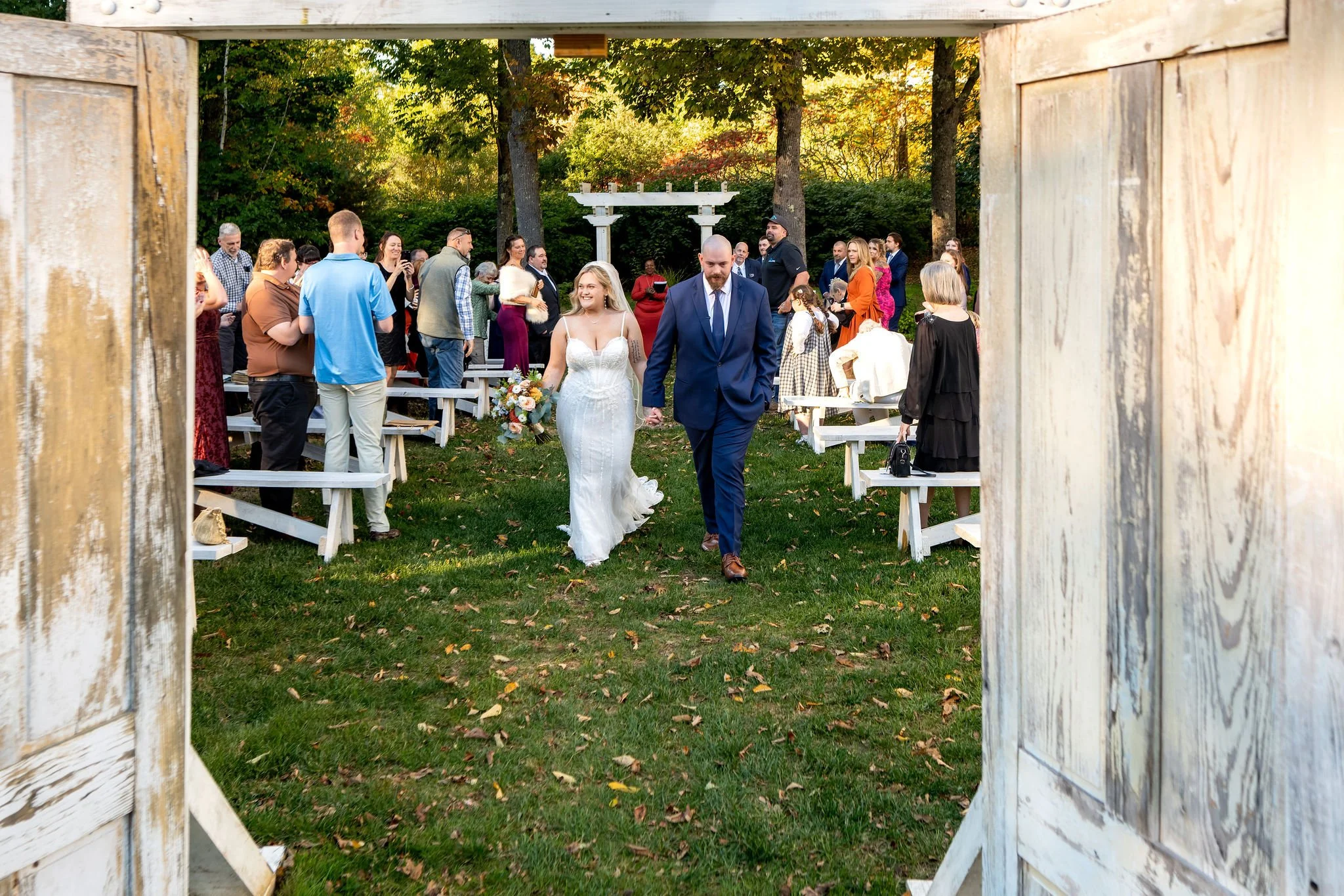 Ellie and her dad walking the aisle seen through the open weathered doors as guests stand to watch at Boothbay Playhouse in Boothbay Maine fall wedding photography VMG Productions