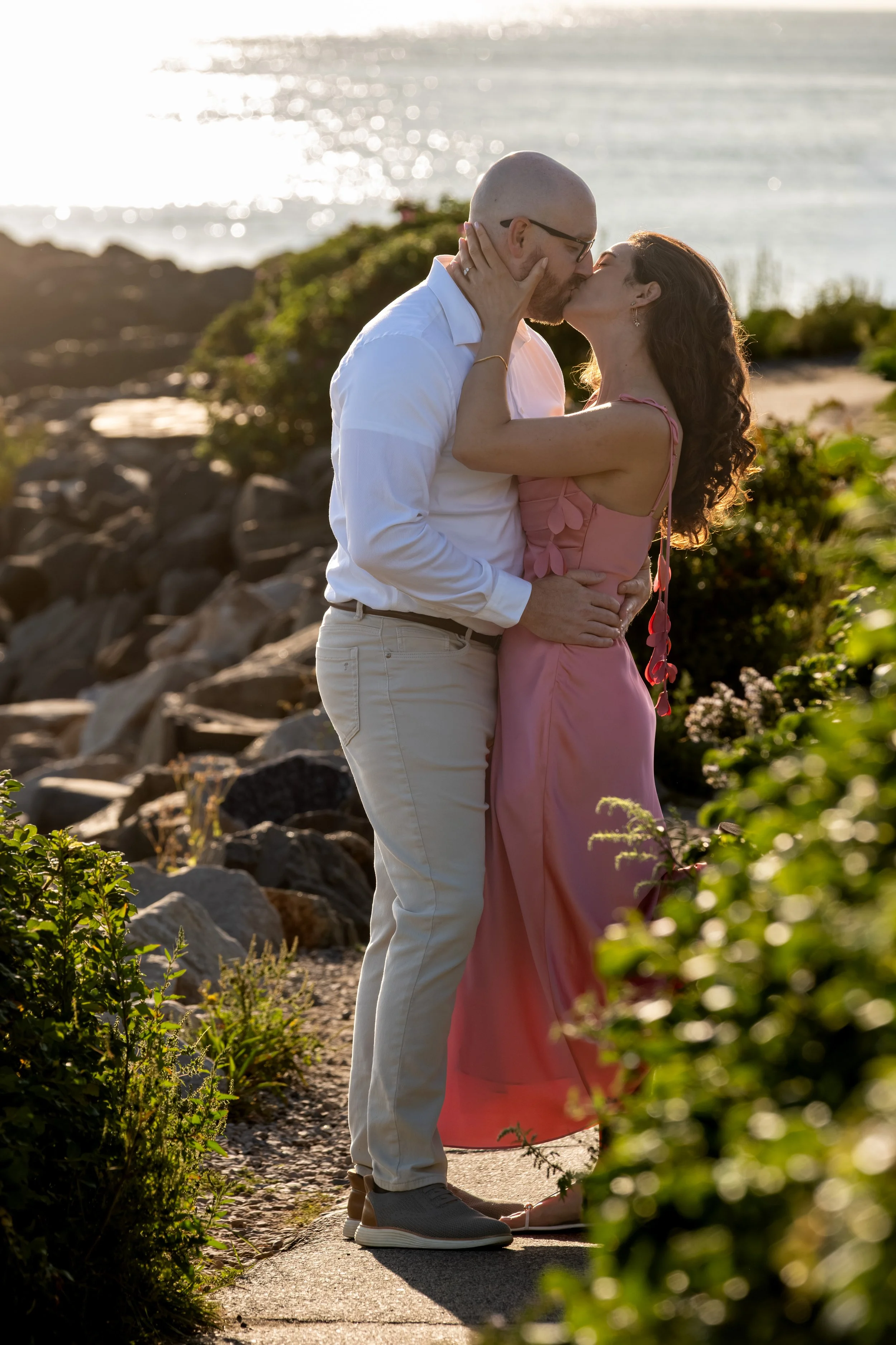 Zack and Helena kissing on the coastal path with the roses framing them and the ocean glittering behind on Marginal Way in Ogunquit Maine sunrise proposal and engagement photography VMG Productions