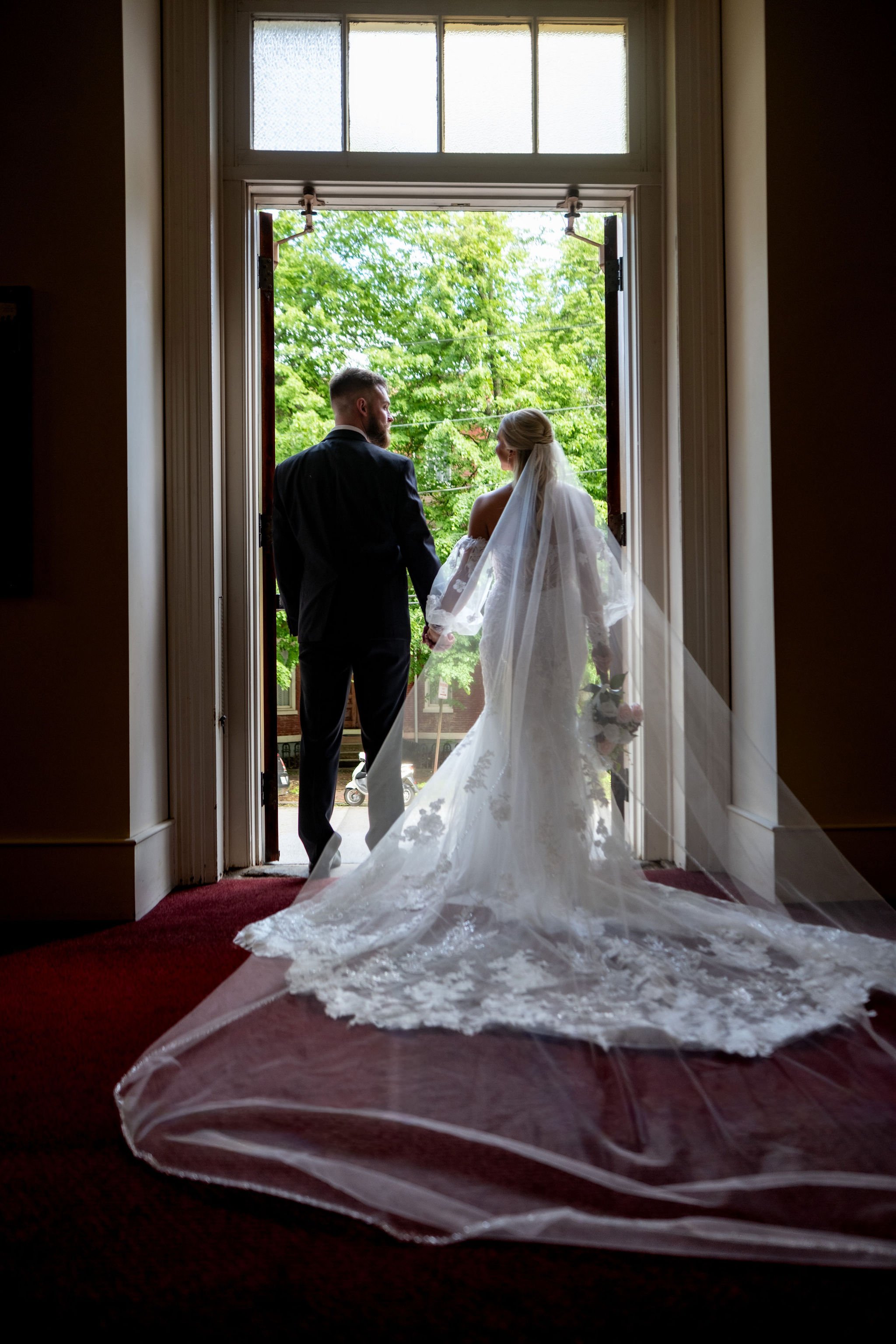 Jeremiah and Vienna couple stands hand in hand inside an open church doorway framed by natural light at Holy Trinity Greek Orthodox Church Portland Maine spring wedding photography VMG Productions
