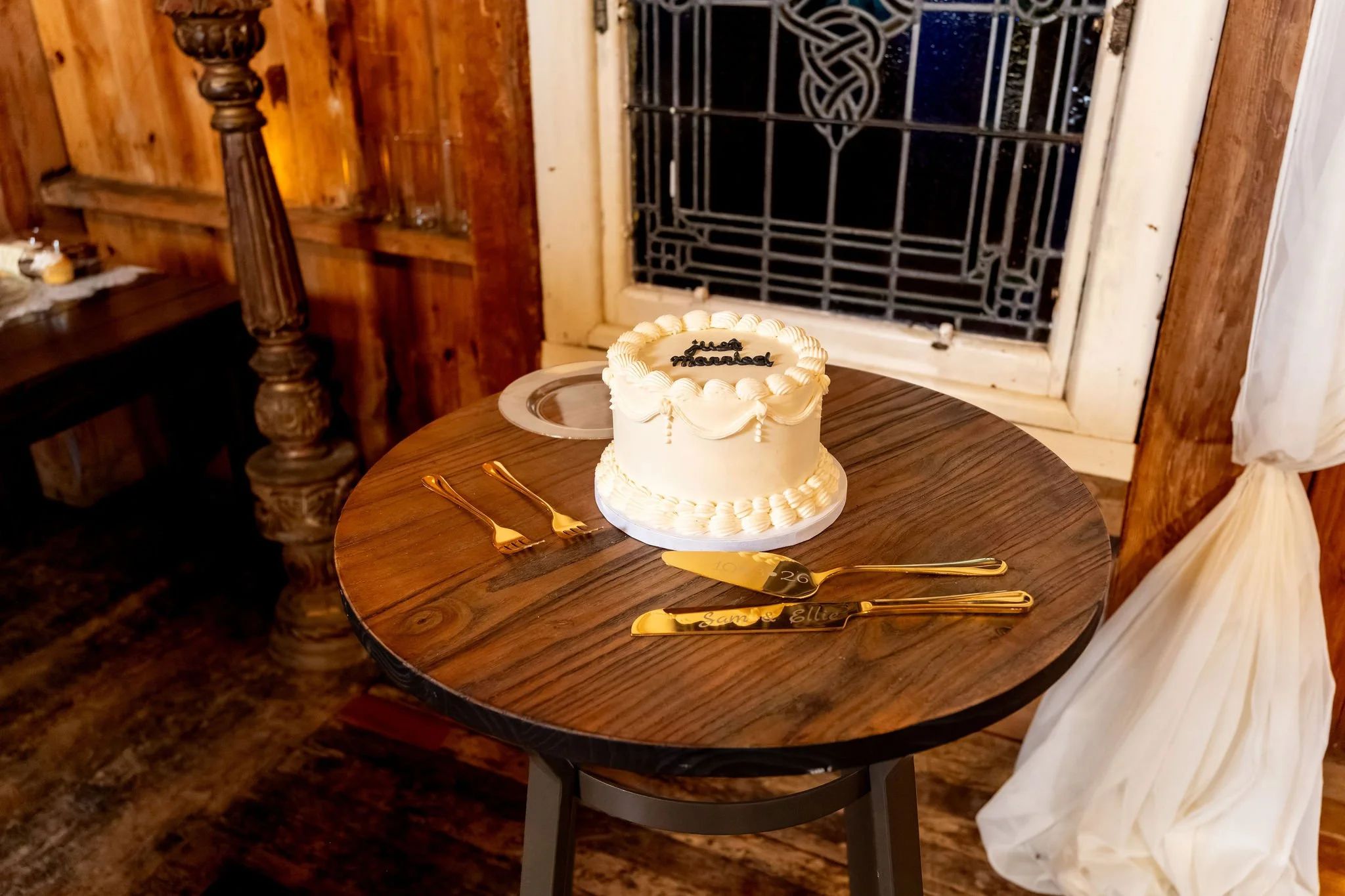 A small white wedding cake on a round wooden table by the stained glass window at Boothbay Playhouse in Boothbay Maine fall wedding photography VMG Productions