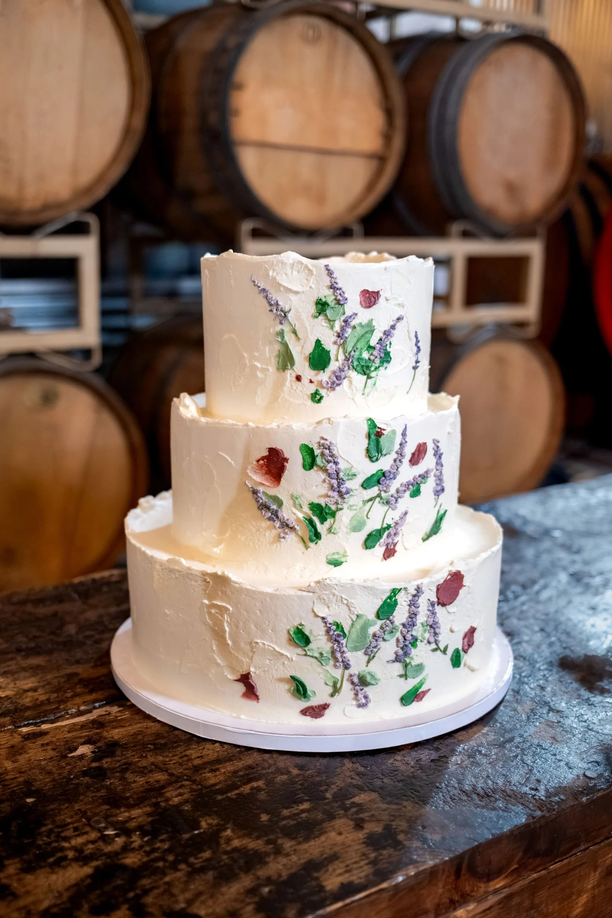 Three tier white wedding cake hand painted with lavender sprigs and petals on a dark wood table at The Cellars at Allagash Brewing Company in Portland Maine fall wedding photography VMG Productions