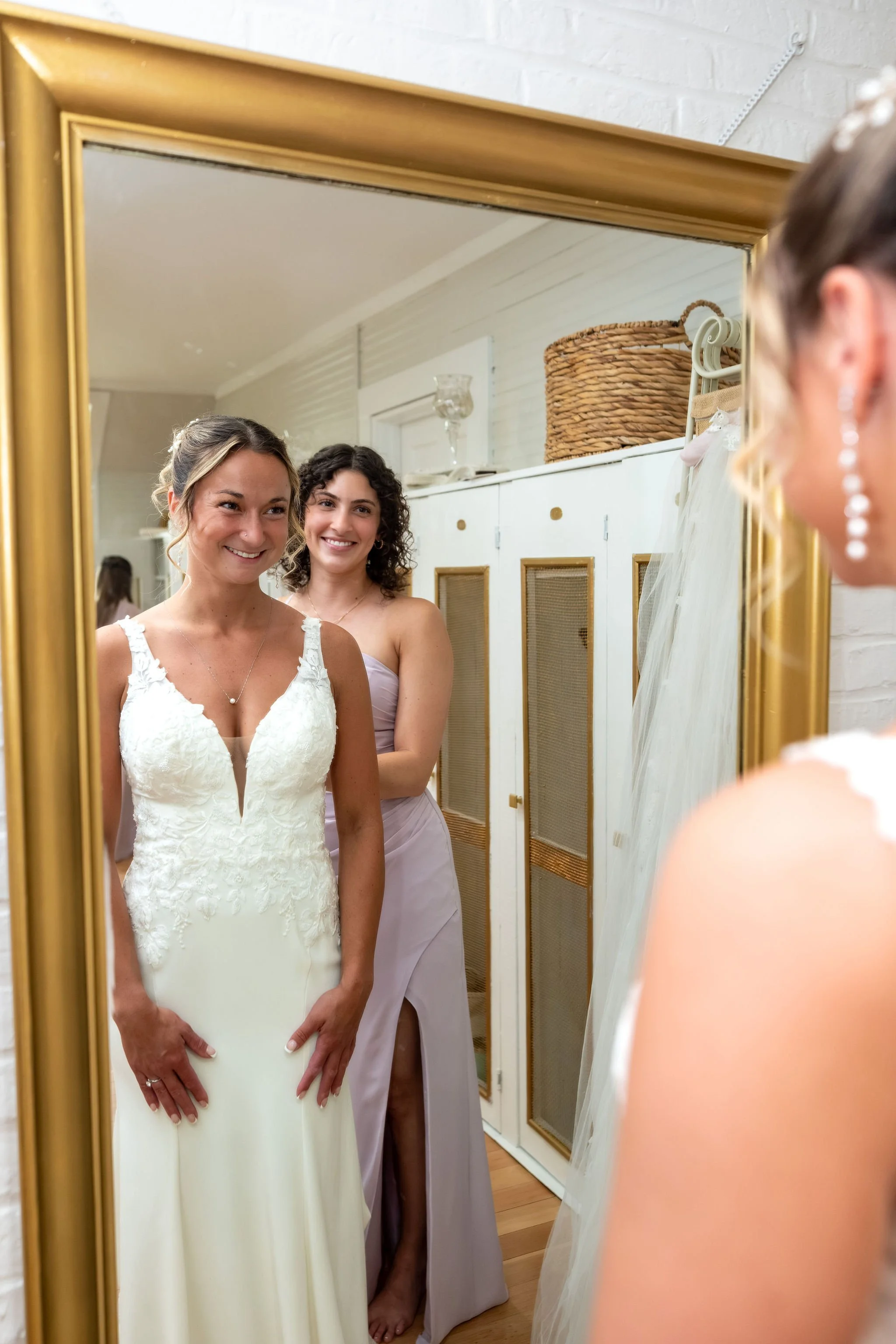 Beth and Cameron bride and a bridesmaid smiling in the mirror while getting ready at Augusta Country Club Manchester Maine spring wedding photography VMG Productions