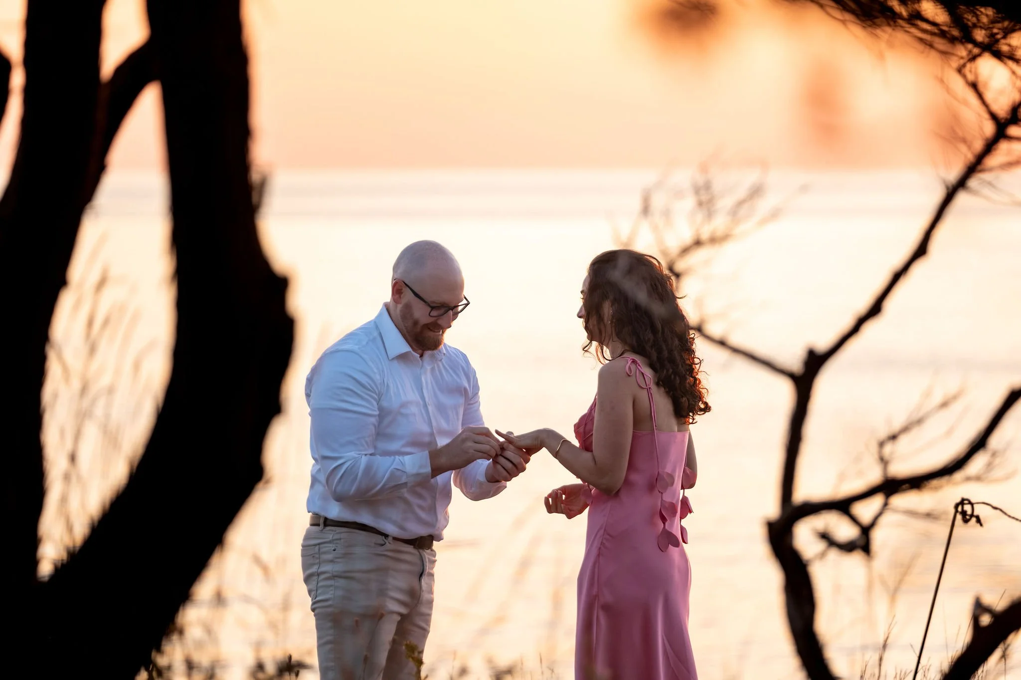 Zack slipping the ring onto Helena's finger with the pink sunrise sky and ocean behind them on Marginal Way in Ogunquit Maine sunrise proposal and engagement photography VMG Productions