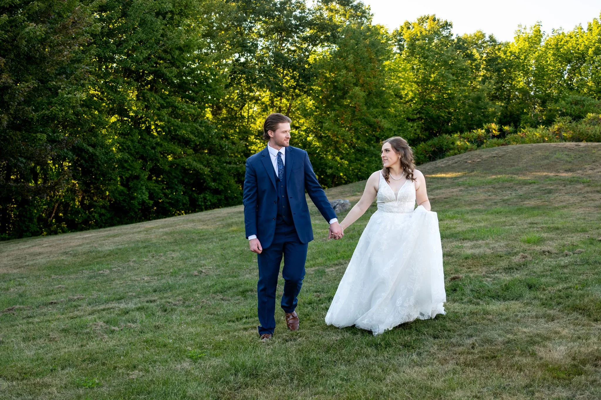 Zachary and Hollie walking hand in hand across the sloping field toward the trees at The Lucerne Inn in Dedham Maine summer wedding photography VMG Productions