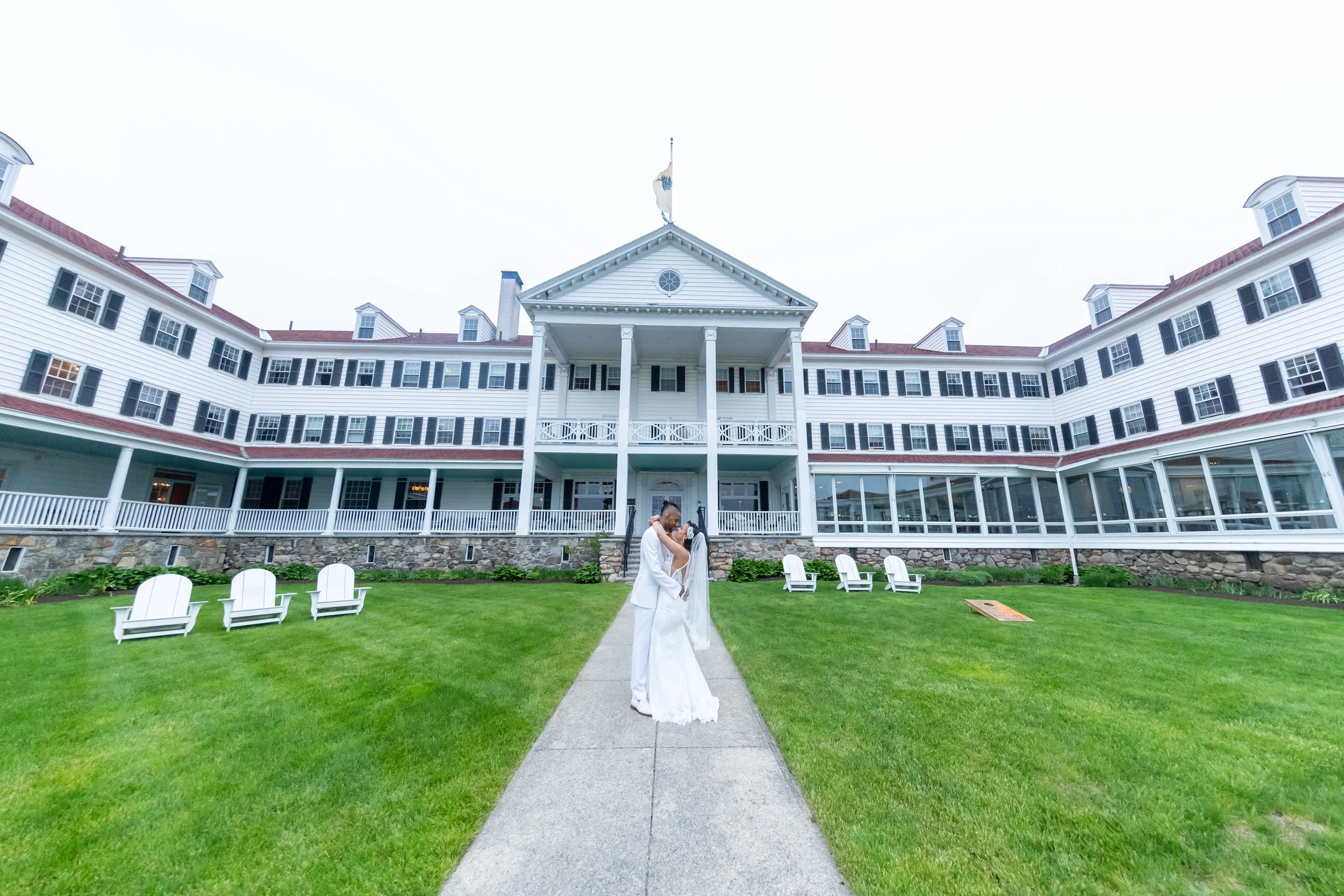 Bride in white lace wedding gown and groom in white suit embracing on center pathway in front of grand white Colonial hotel facade with Adirondack chairs on lawn at The Colony Hotel Kennebunkport Maine May 2022