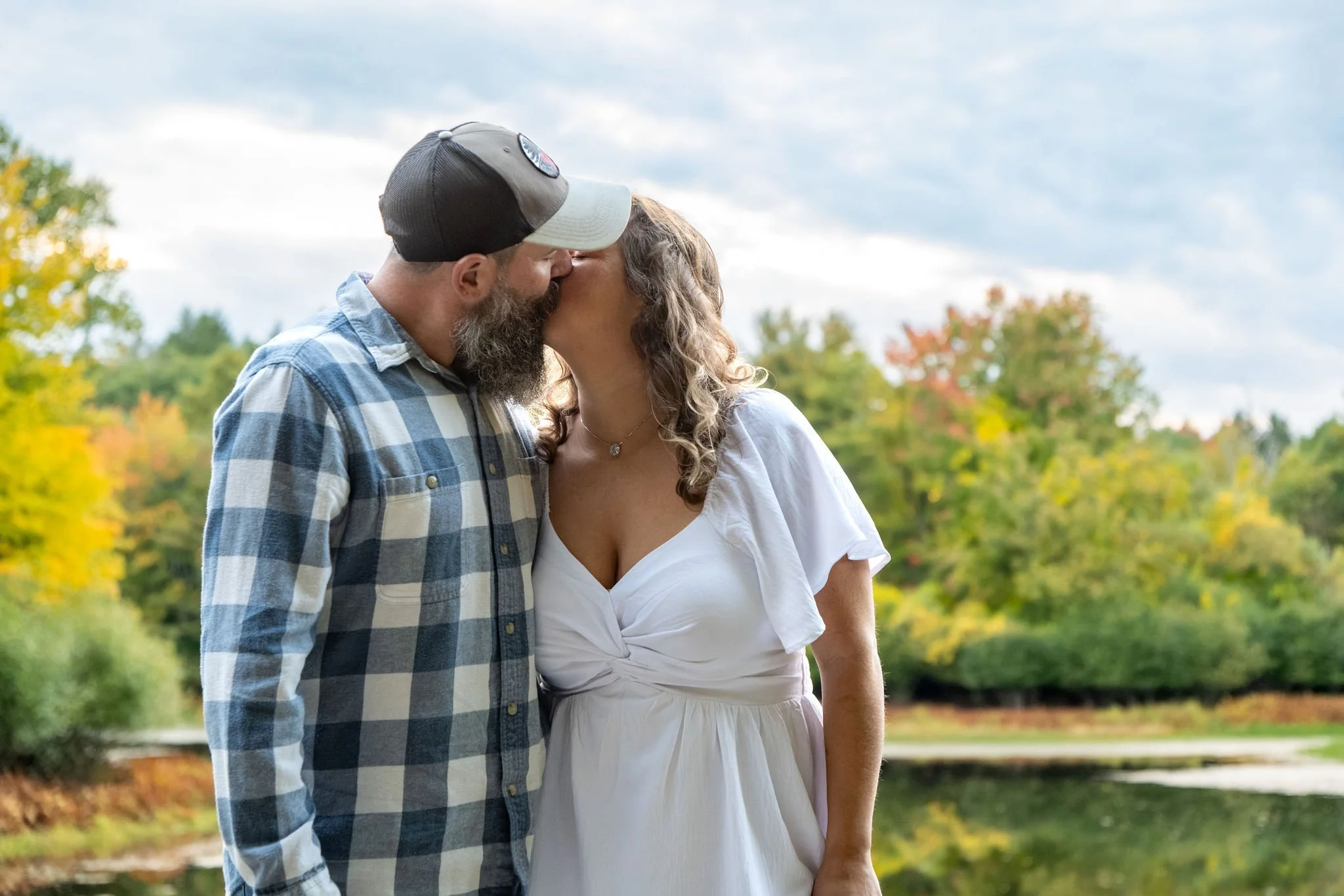 Alison and Dave kissing under a bright sky with yellow and orange trees along the far shore of the pond at Vaughan Woods in Hallowell Maine fall engagement photography VMG Productions