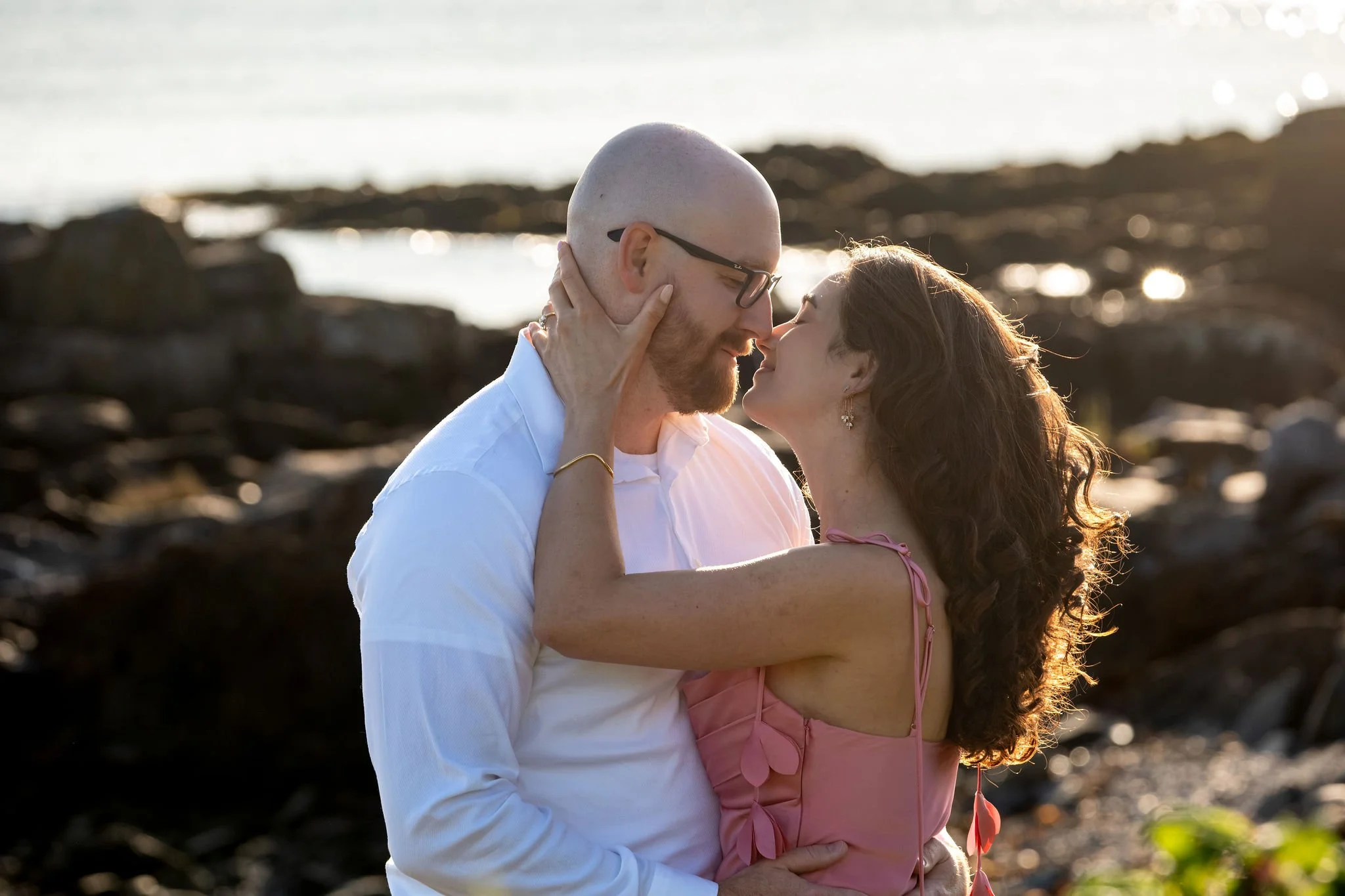 Zack cradling Helena's face as they kiss with the golden light glowing through her hair on Marginal Way in Ogunquit Maine sunrise proposal and engagement photography VMG Productions