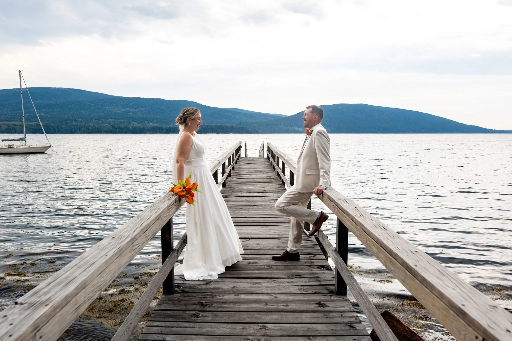 Alli and Adam Baudoin leaning on opposite railings of the wooden dock over Somes Sound with the Acadia hills behind them in Mount Desert Maine spring wedding photography VMG Productions