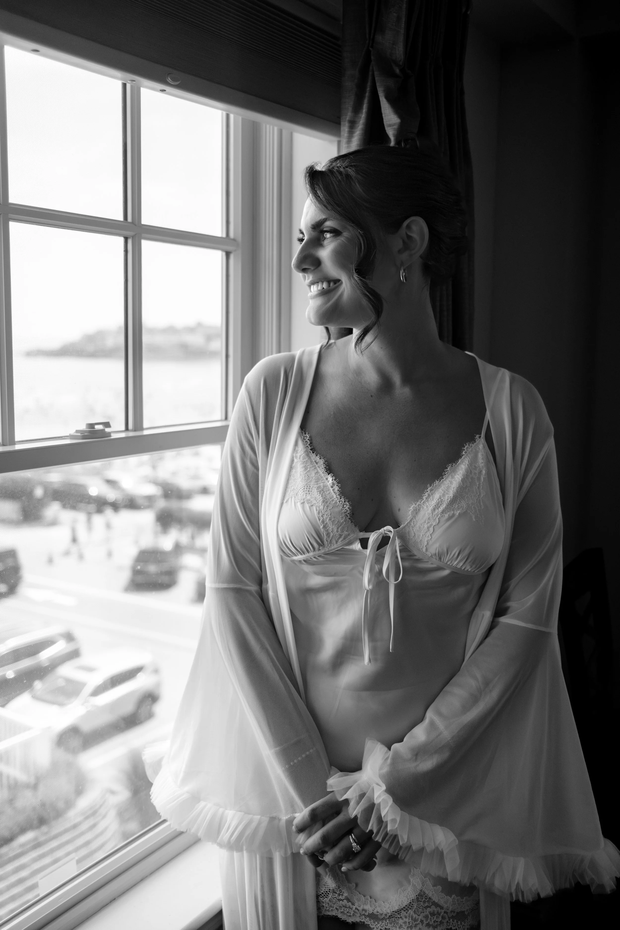 Caroline Pirolli smiling at the window in her robe with the beach visible below in black and white at Union Bluff Hotel in York Maine wedding photography VMG Productions