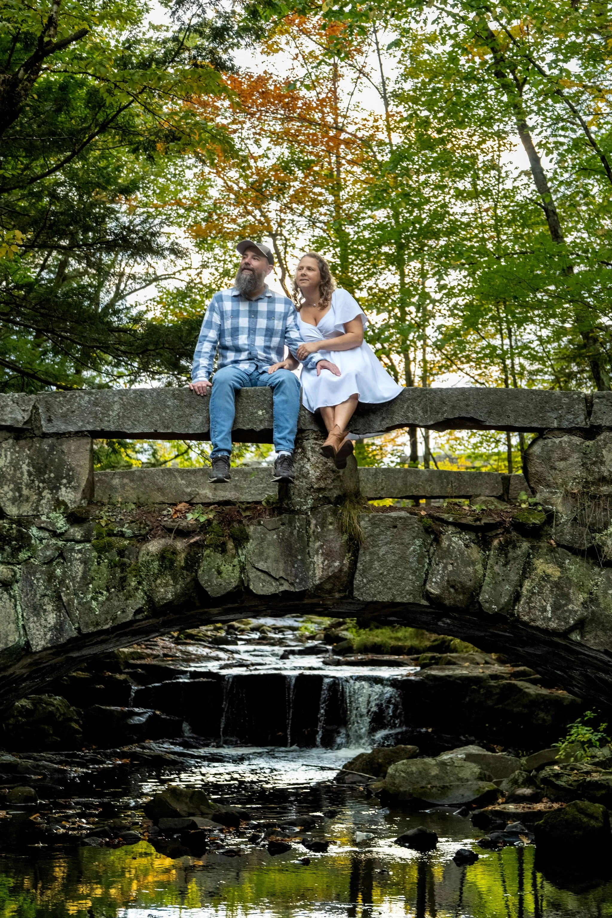 Alison and Dave sitting on the bridge wall with their boots hanging over the arch at Vaughan Woods in Hallowell Maine fall engagement photography VMG Productions