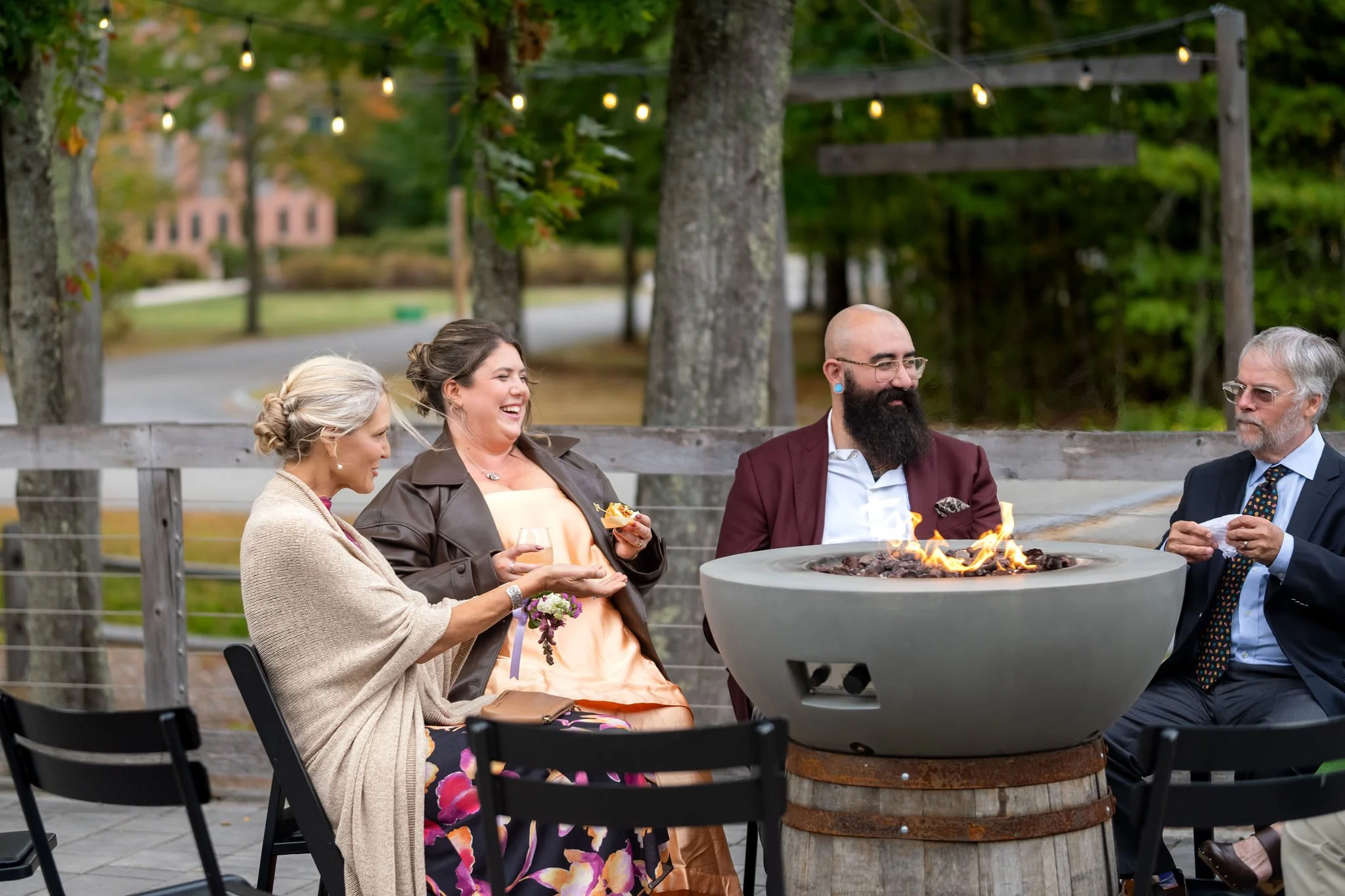 Guests laughing around a gas fire pit on the patio under string lights at The Cellars at Allagash Brewing Company in Portland Maine fall wedding photography VMG Productions