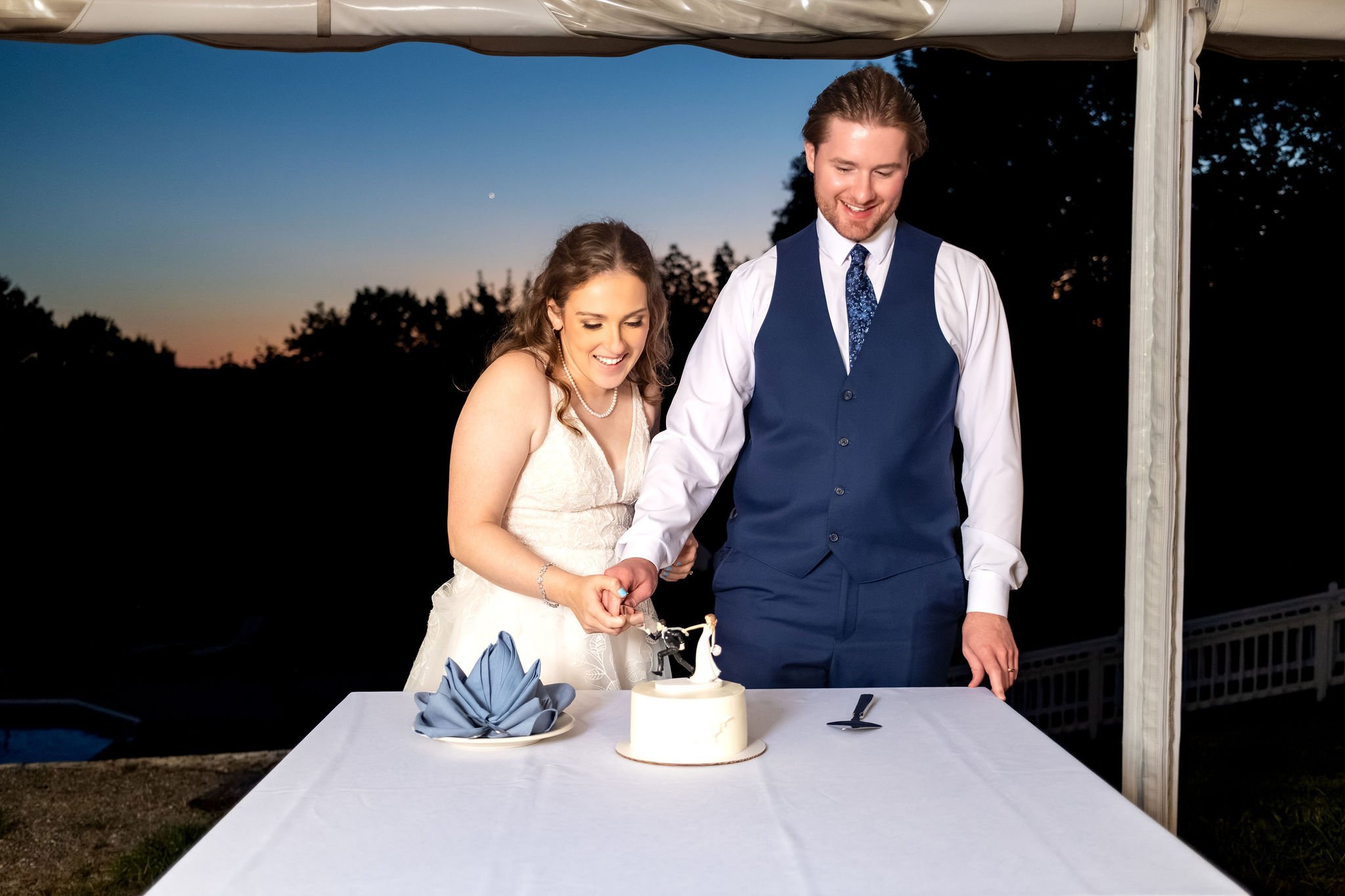 Zachary and Hollie cutting their cake together outside at dusk with the last of the sunset behind them at The Lucerne Inn in Dedham Maine summer wedding photography VMG Productions