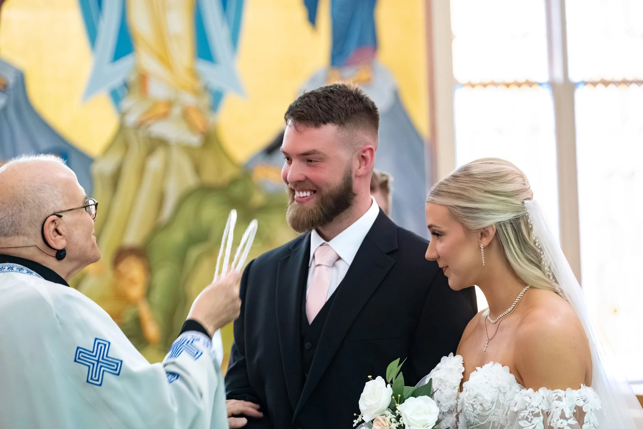 Jeremiah and Vienna the priest presents ceremonial crowns while the couple smiles during a key Orthodox wedding tradition at Holy Trinity Greek Orthodox Church Portland Maine spring wedding photography VMG Productions