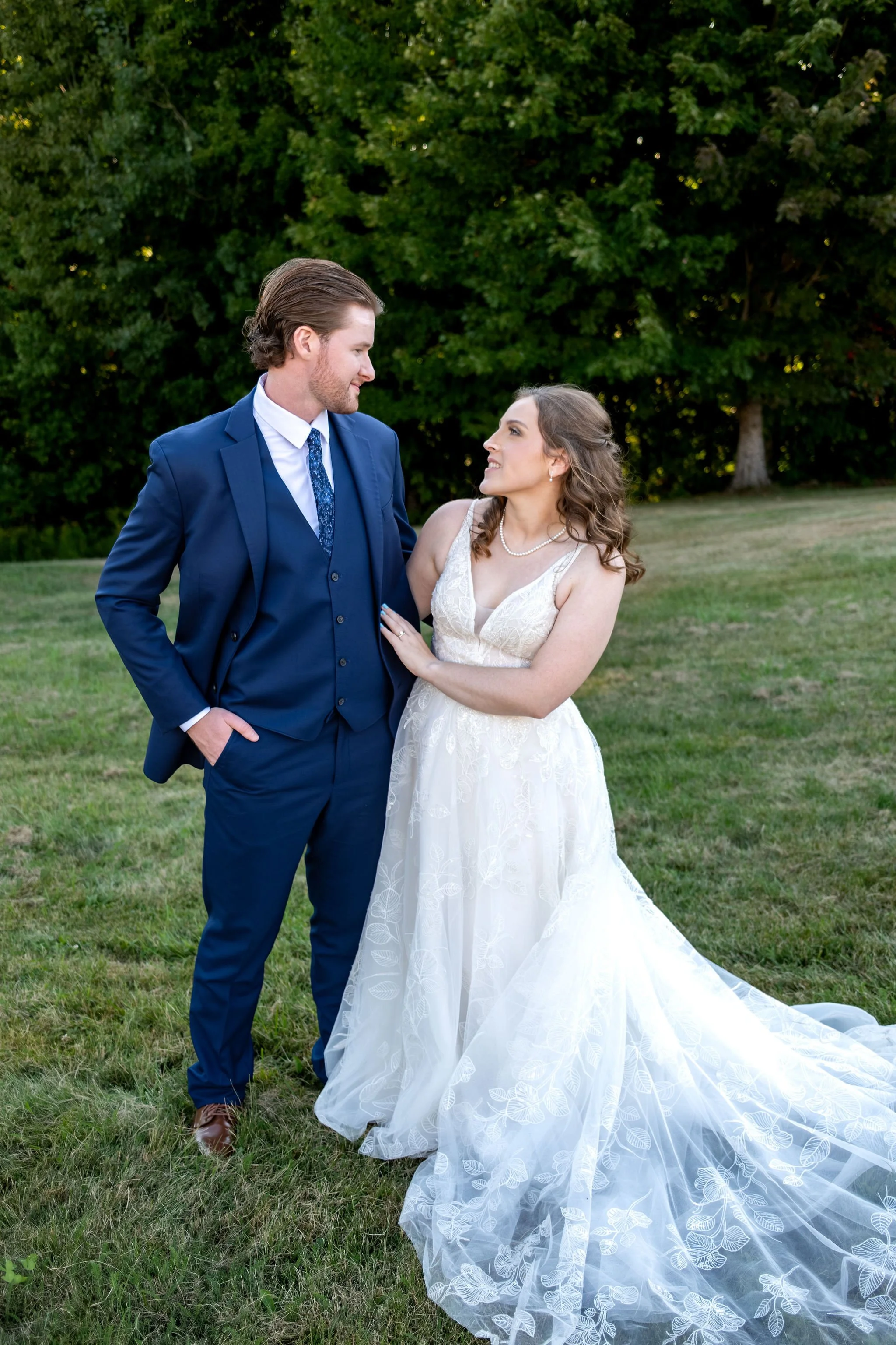 Zachary and Hollie looking at each other on the lawn with her lace train spread across the grass at The Lucerne Inn in Dedham Maine summer wedding photography VMG Productions