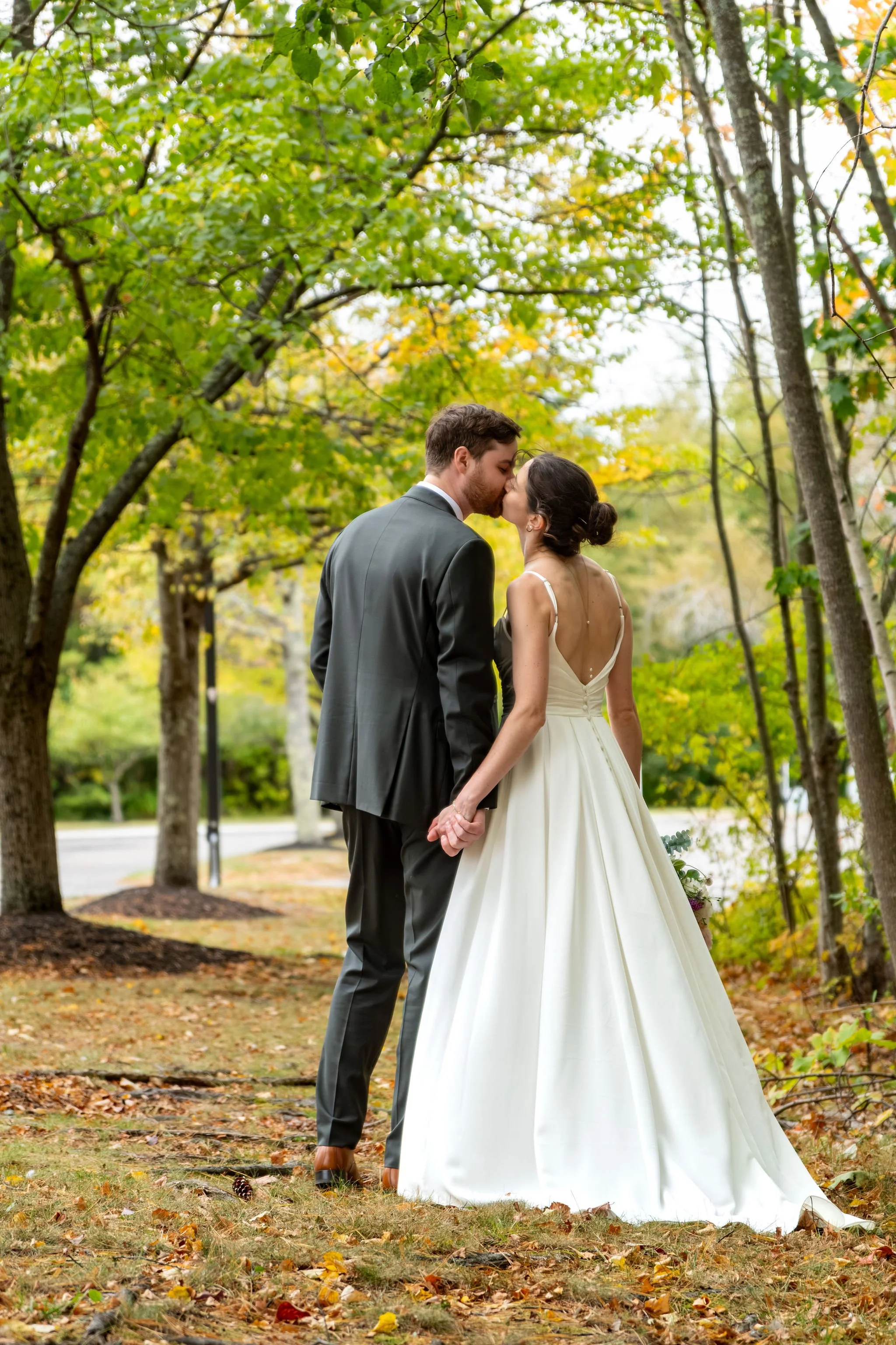Victoria and Jack kissing from behind in the trees with her open back gown and train on the leaves at The Cellars at Allagash Brewing Company in Portland Maine fall wedding photography VMG Productions