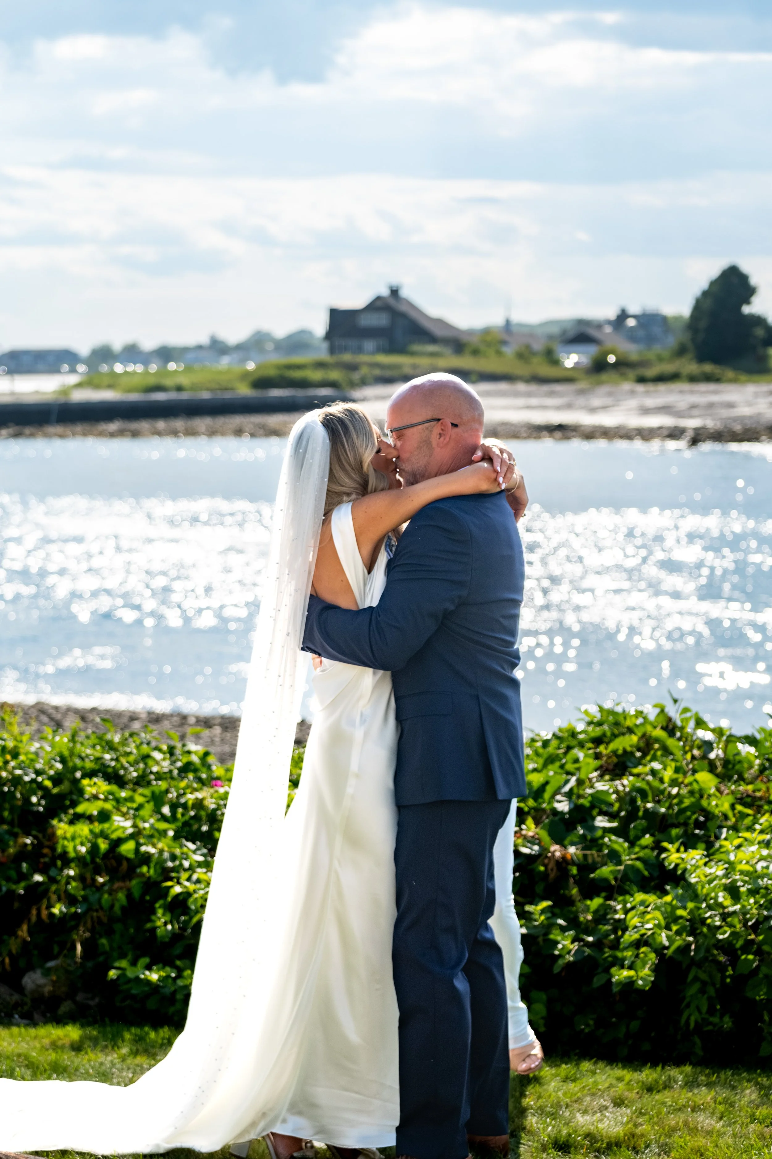 Ashley and Marc Draper sharing their first kiss as a married couple with the water sparkling behind them at The Breakwater Inn in Kennebunkport Maine summer wedding photography VMG Productions