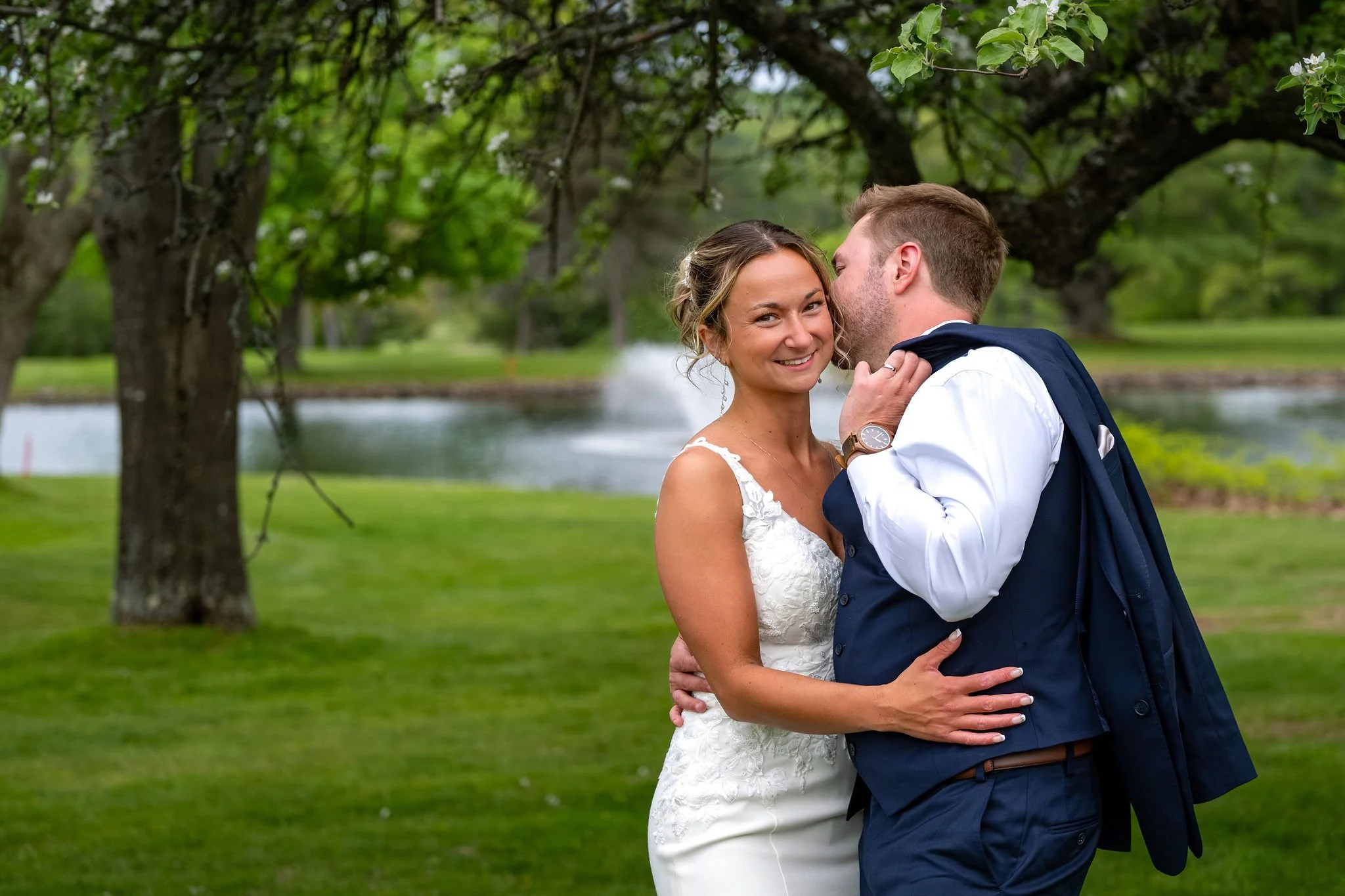 Beth and Cameron smiling at the camera as the groom leans in beneath a flowering tree at Augusta Country Club Manchester Maine spring wedding photography VMG Productions