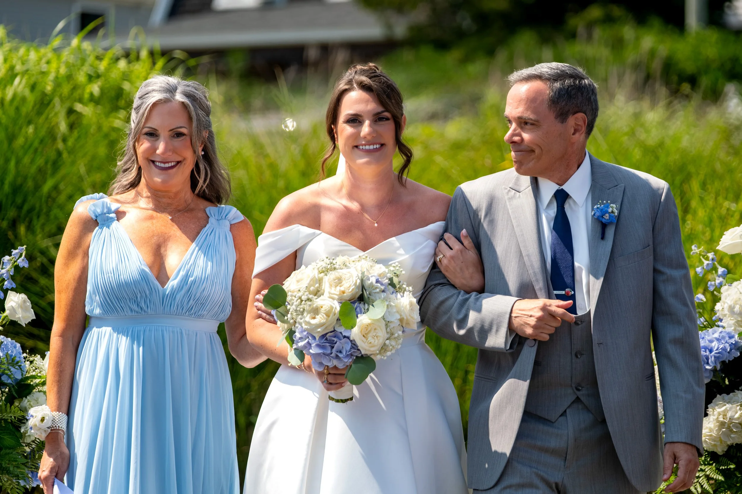 Caroline Pirolli walking the aisle with both parents beside her through the blue hydrangea arrangements at Union Bluff Hotel in York Maine wedding photography VMG Productions